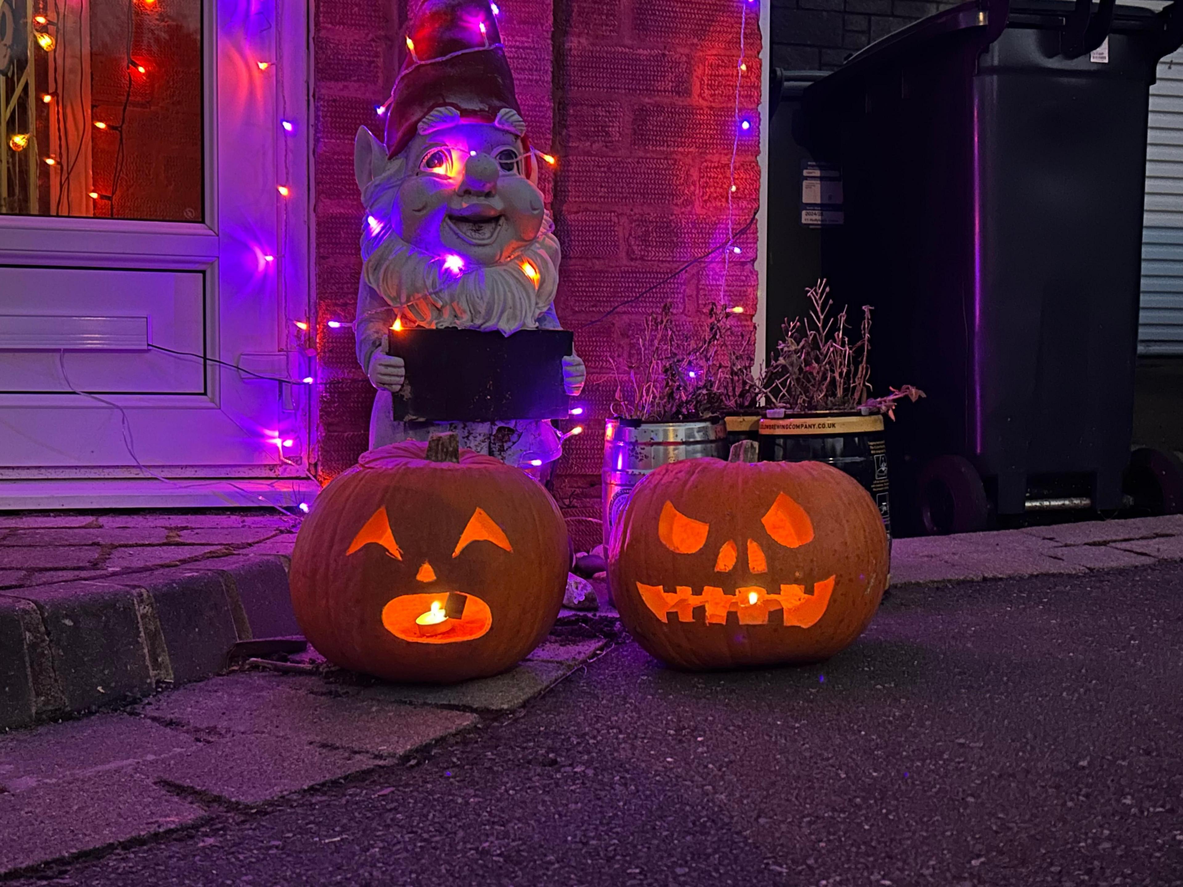 Two carved pumpkins by a doorstep. Behind them, a garden gnome and front door are decorated with coloured lights.  