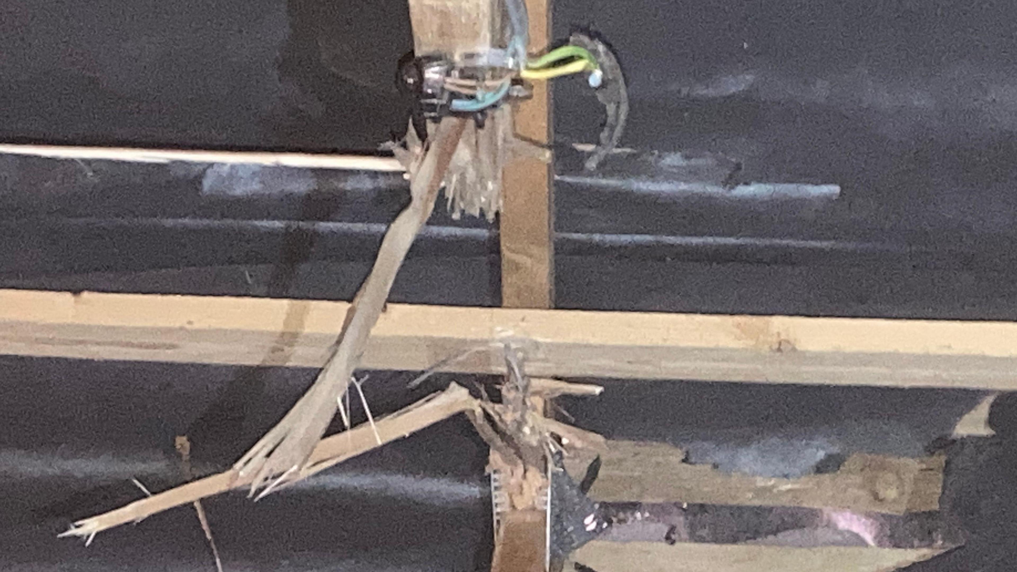 Splintered roof beams and damaged black insulation material. Some wires are hanging down  exposed from the ceiling.