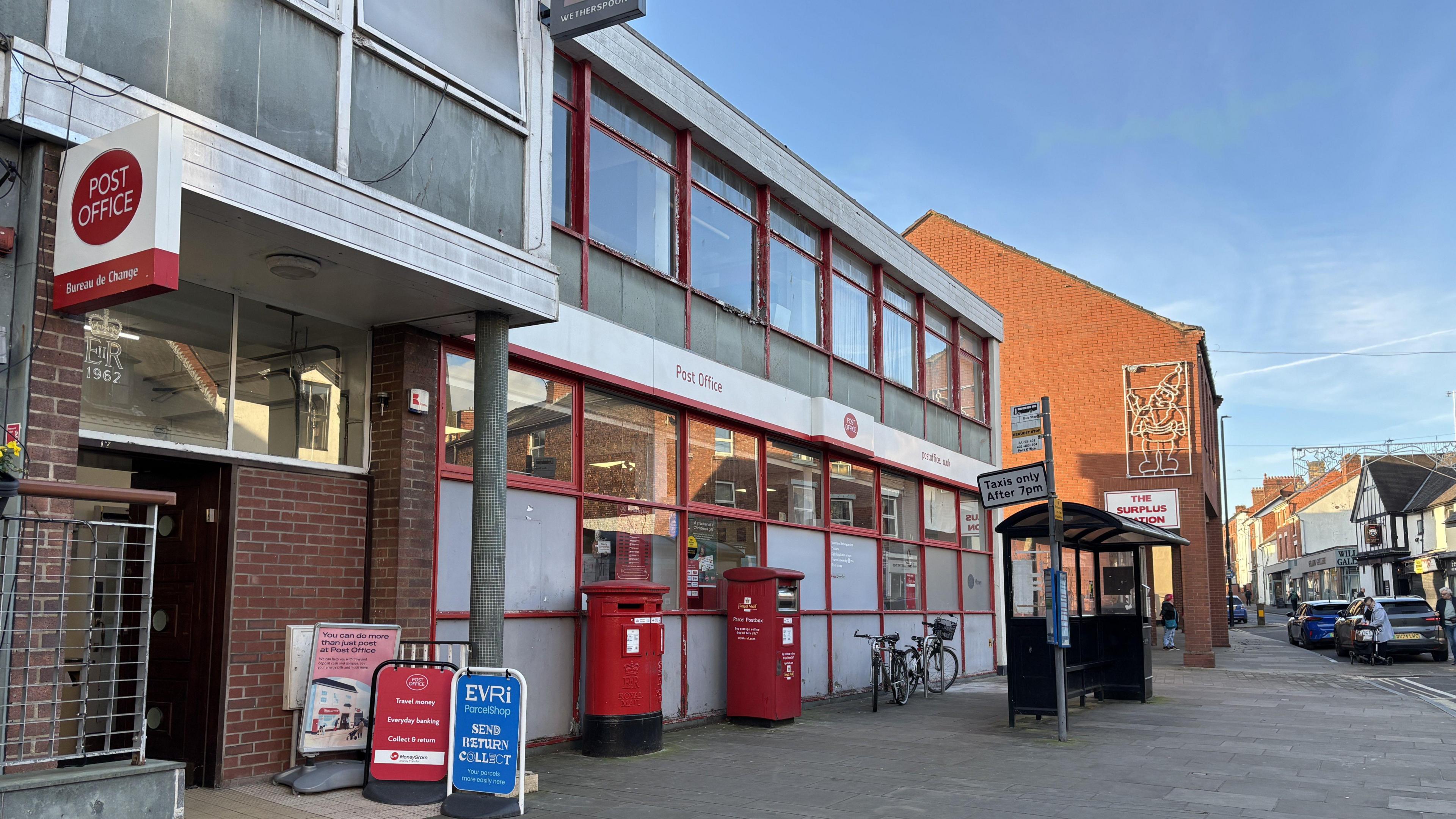 The outside of Oswestry Post Office. The building has lots of windows with red panelling. There is a white sign that reads "Post Office". The entrance is red brick and there are two red post boxes outside for letters and parcels.