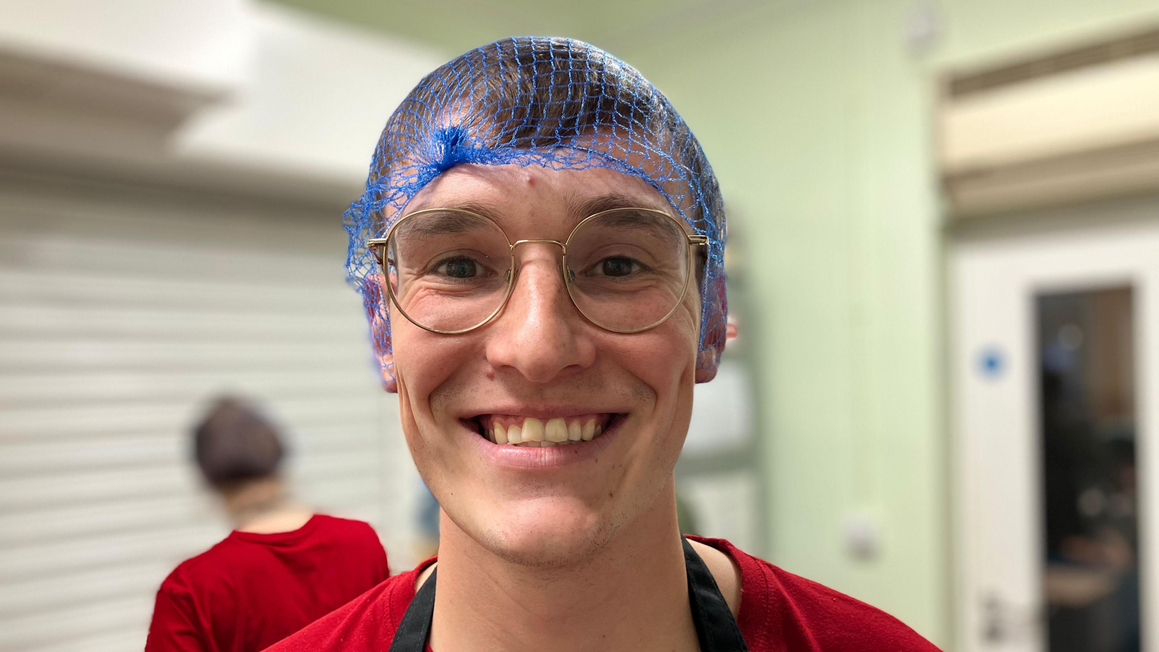 A smiling man with round-framed glasses is wearing a blue hair net, red T-shirt and a black apron on top.