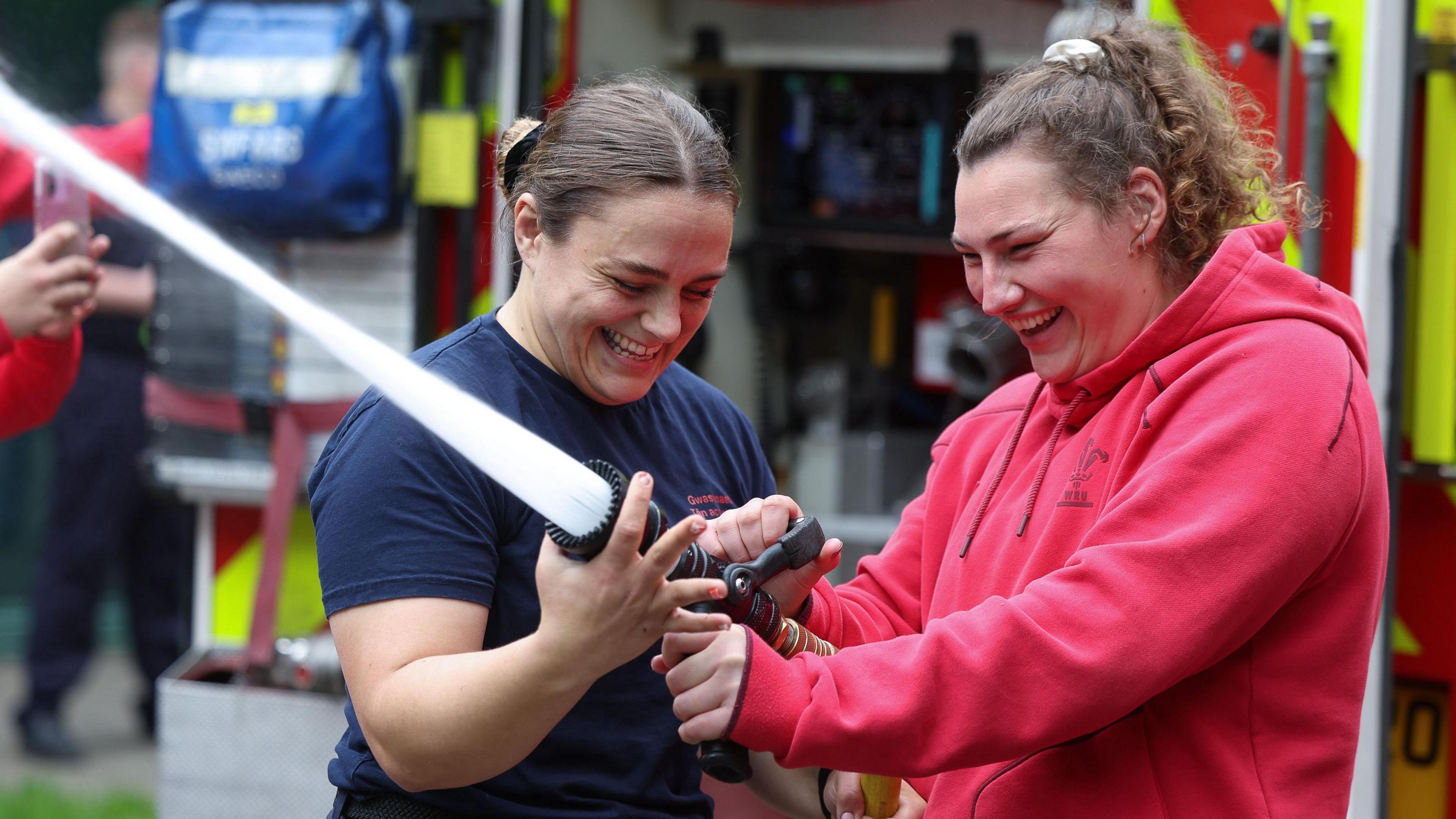 Firefighter Jenni Scoble shows Wales team-mate Gwenllian Pyrs how to use a hose gushing water.