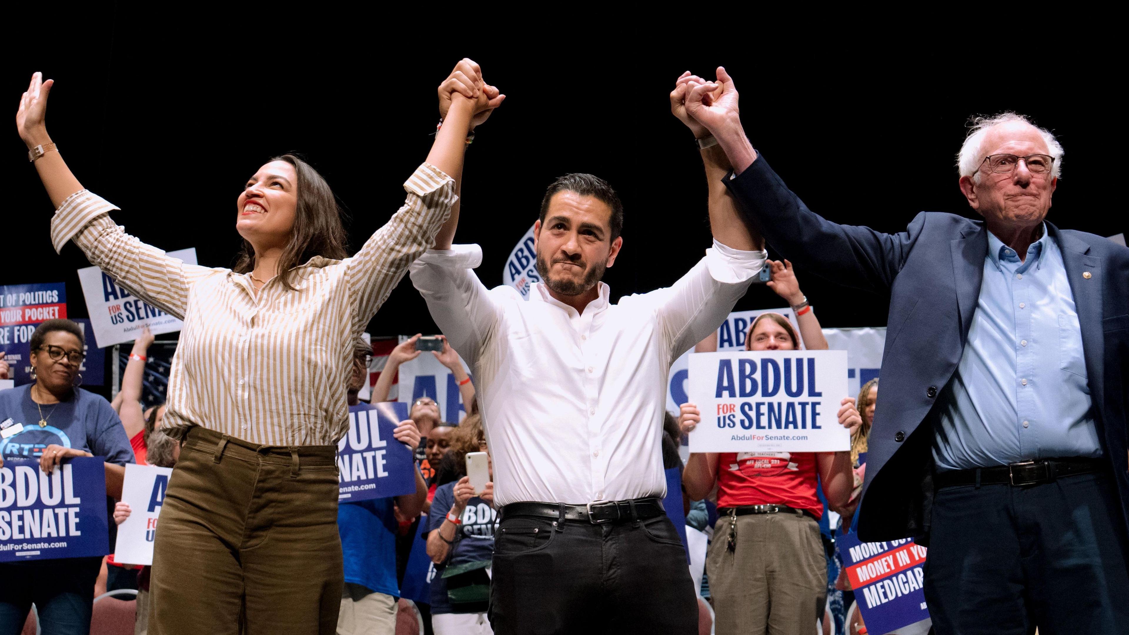 Alexandria Ocasio-Cortez, a Democrat from New York, Abdul El-Sayed, US Democratic Senate candidate from Michigan, and Senator Bernie Sanders all hold hands aloft in front of campaigners holding signs in support of El-Sayed