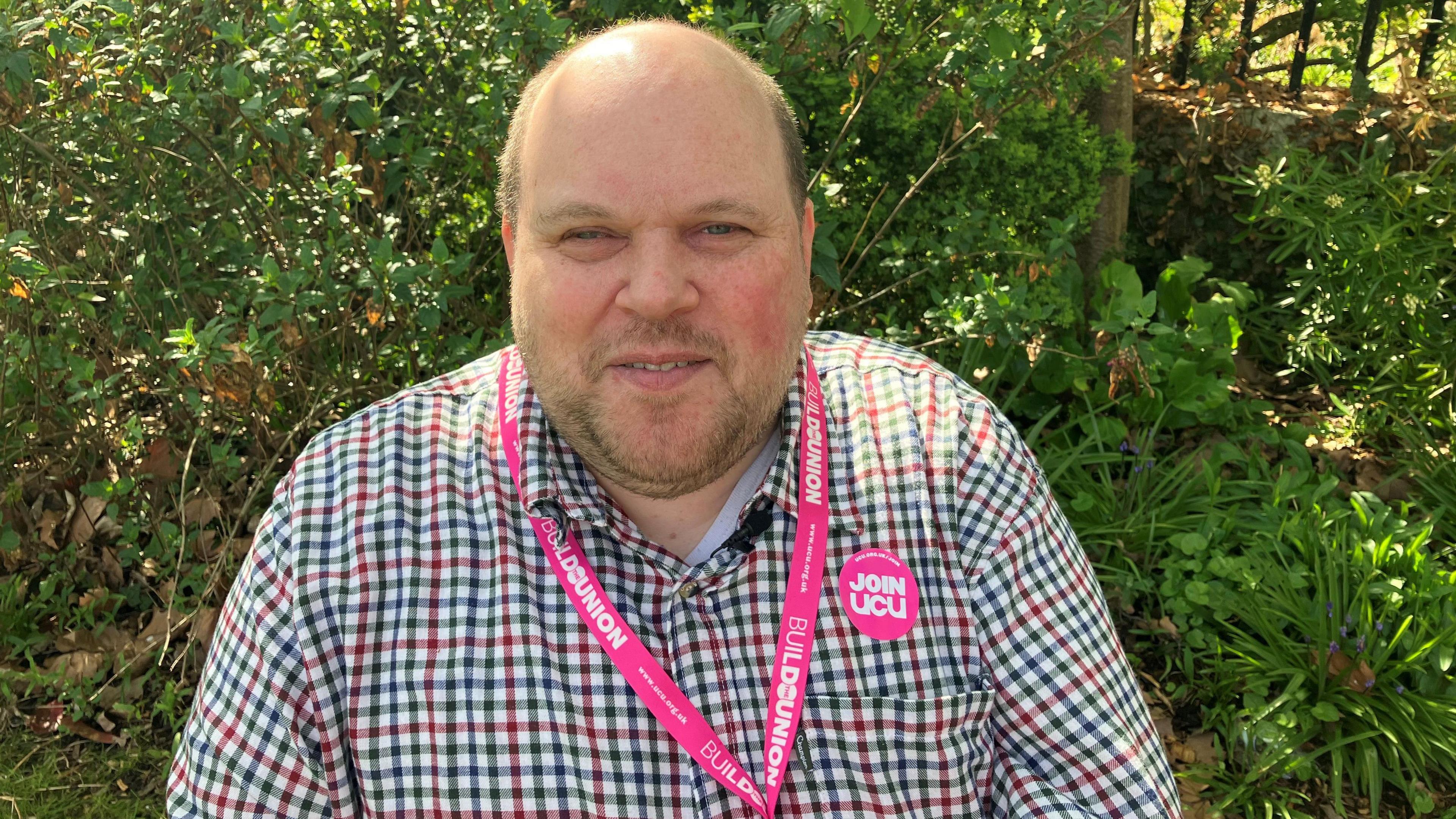 Craig Potter, wearing a black and red checked shirt, sits in a garden. He is wearing a pink lanyard and pink sticker from the University and Colleges Union. 