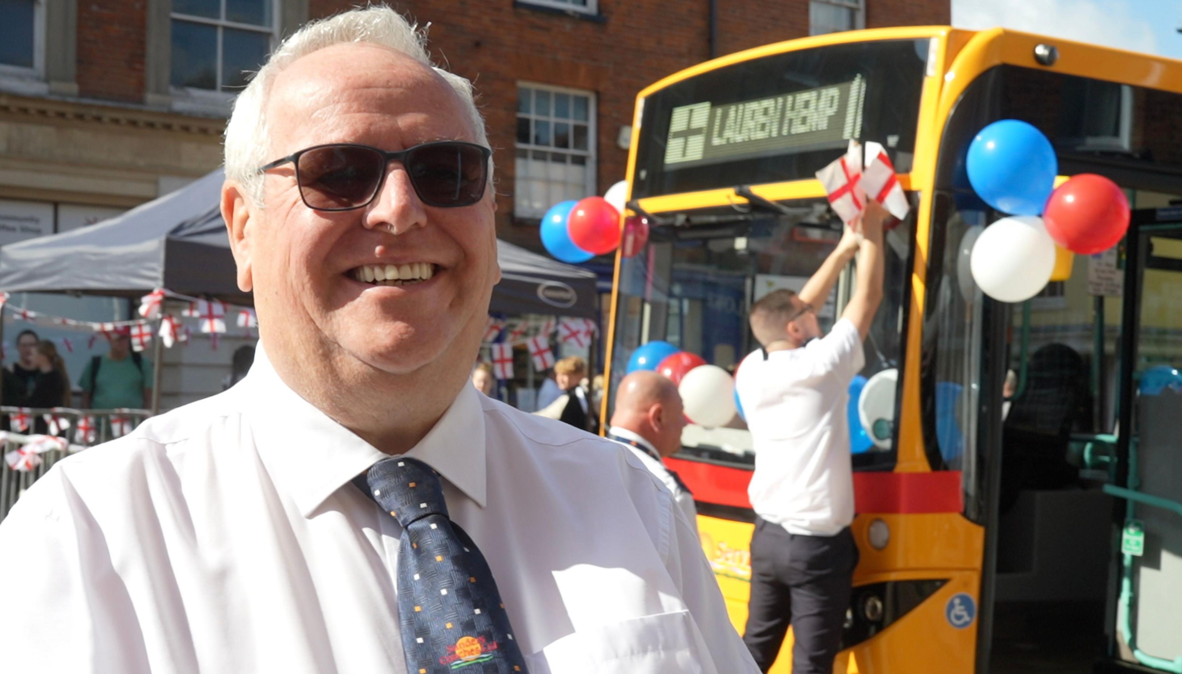 A man with white hair wearing a white shirt and blue tie is smiling and wearing sunglasses as he stands in front of a yellow bus which has balloons and England flags decorating it.