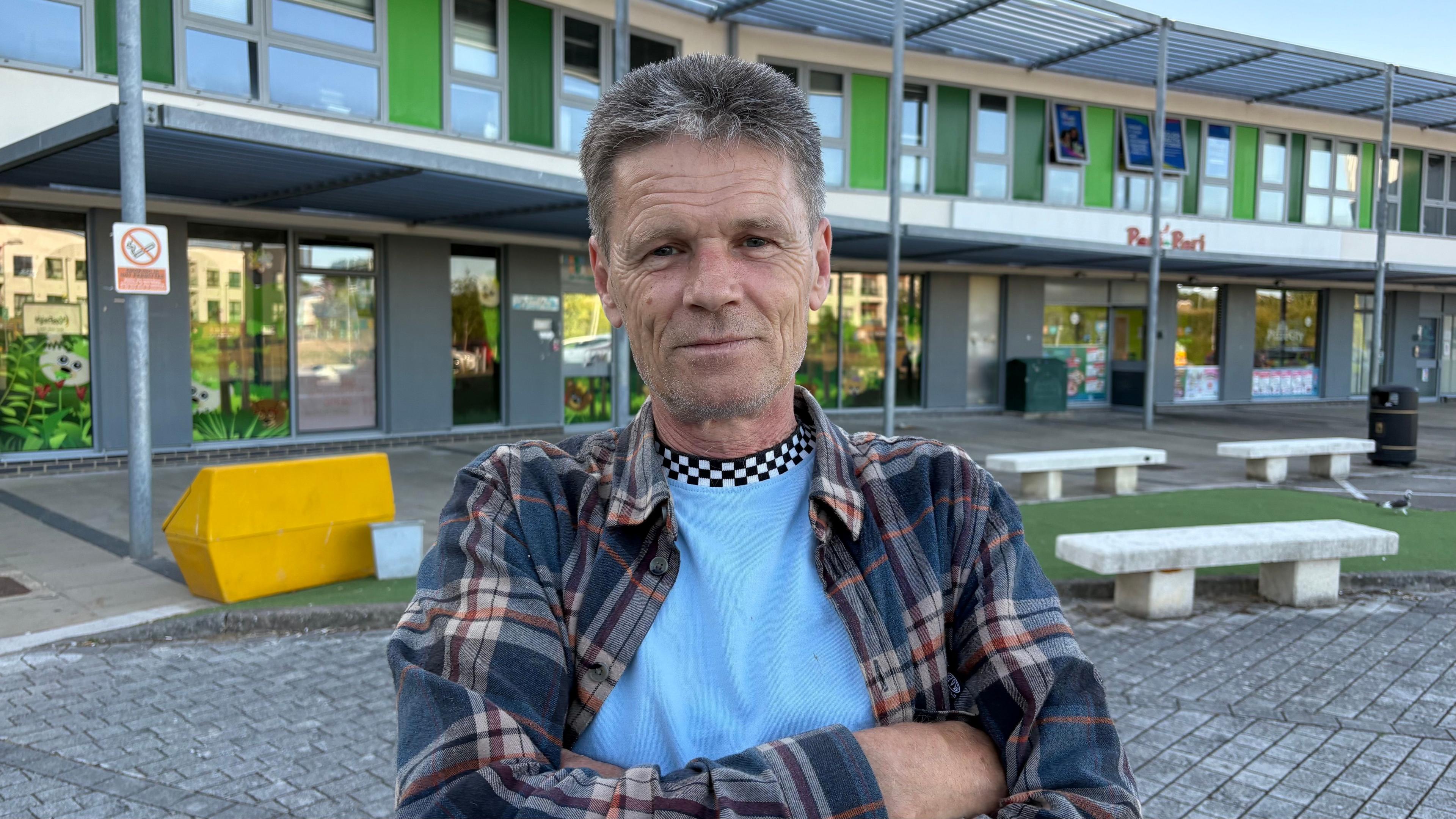 Glenn Jenkins looking directly down the camera. He is wearing an unbuttoned red and blue checked shirt, over a light blue T-shirt that has a black and white checked collar. He has short, greying hair, and his arms are folded. In the background is what looks like a parade of shops, with a cobbled area in the foreground.