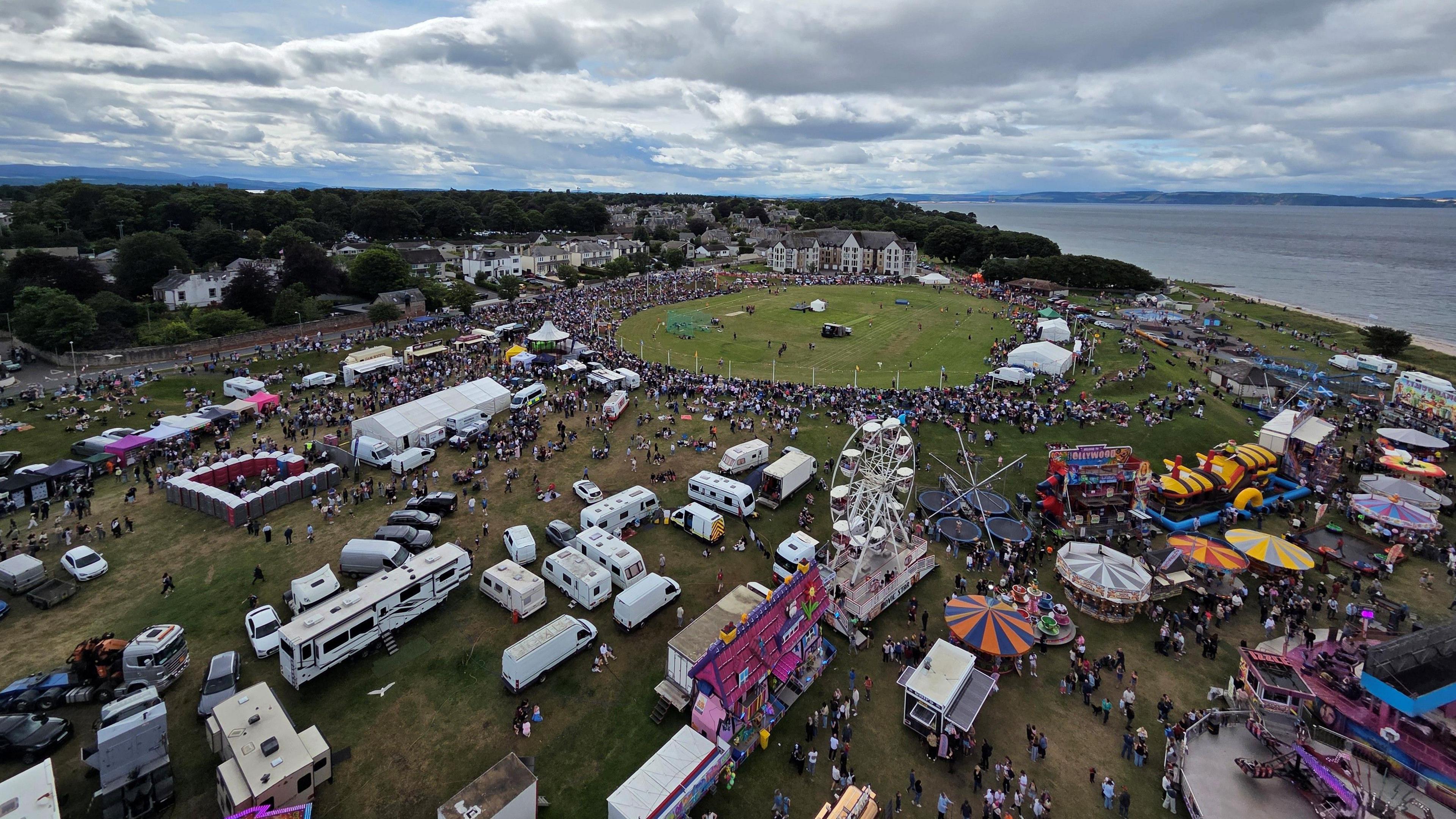 An aerial view of a highland games showing a parade ring, hundreds of people, and fairground rides.