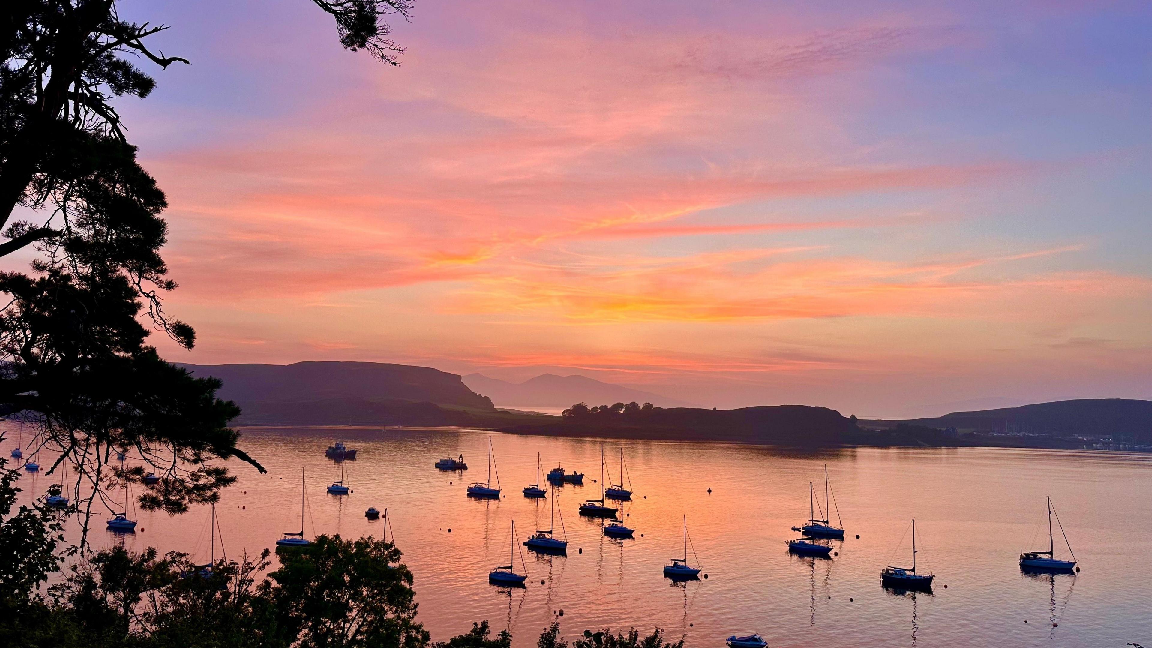 Pink, purple, orange sunset over the water in Oban. The harbour is full of anchored sailing boats.