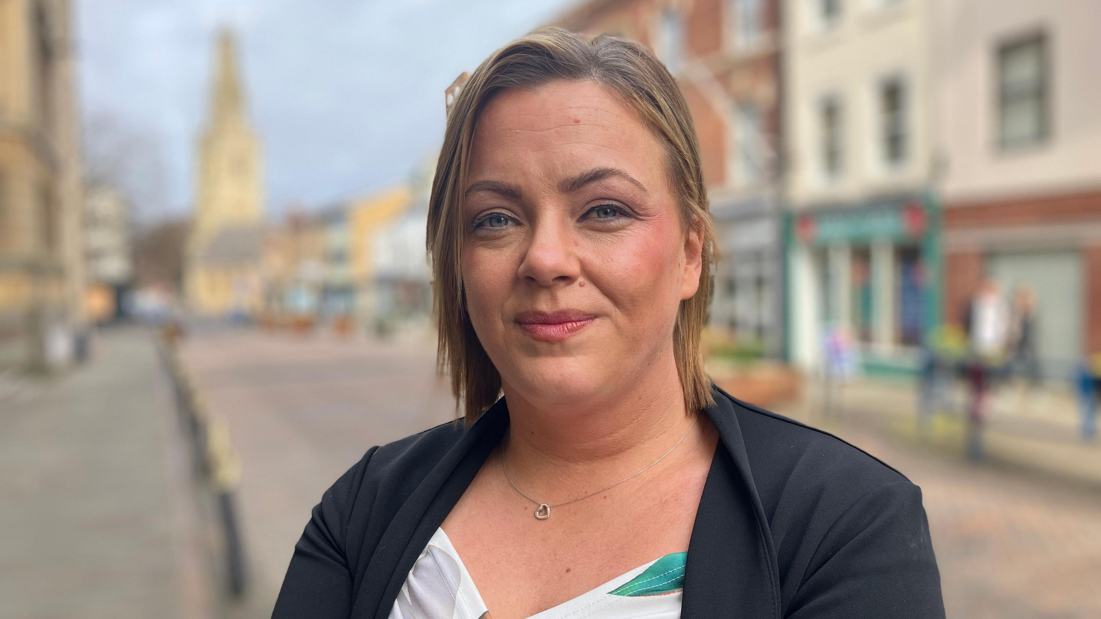 Kate Yhnell, a woman who looks to be in her 40s with a short, dark blonde bob and a black jacket on, with a blue and white top, smiles as she stands in Gloucester City Centre. Blurred shops, businesses and a church can be seen behind her.