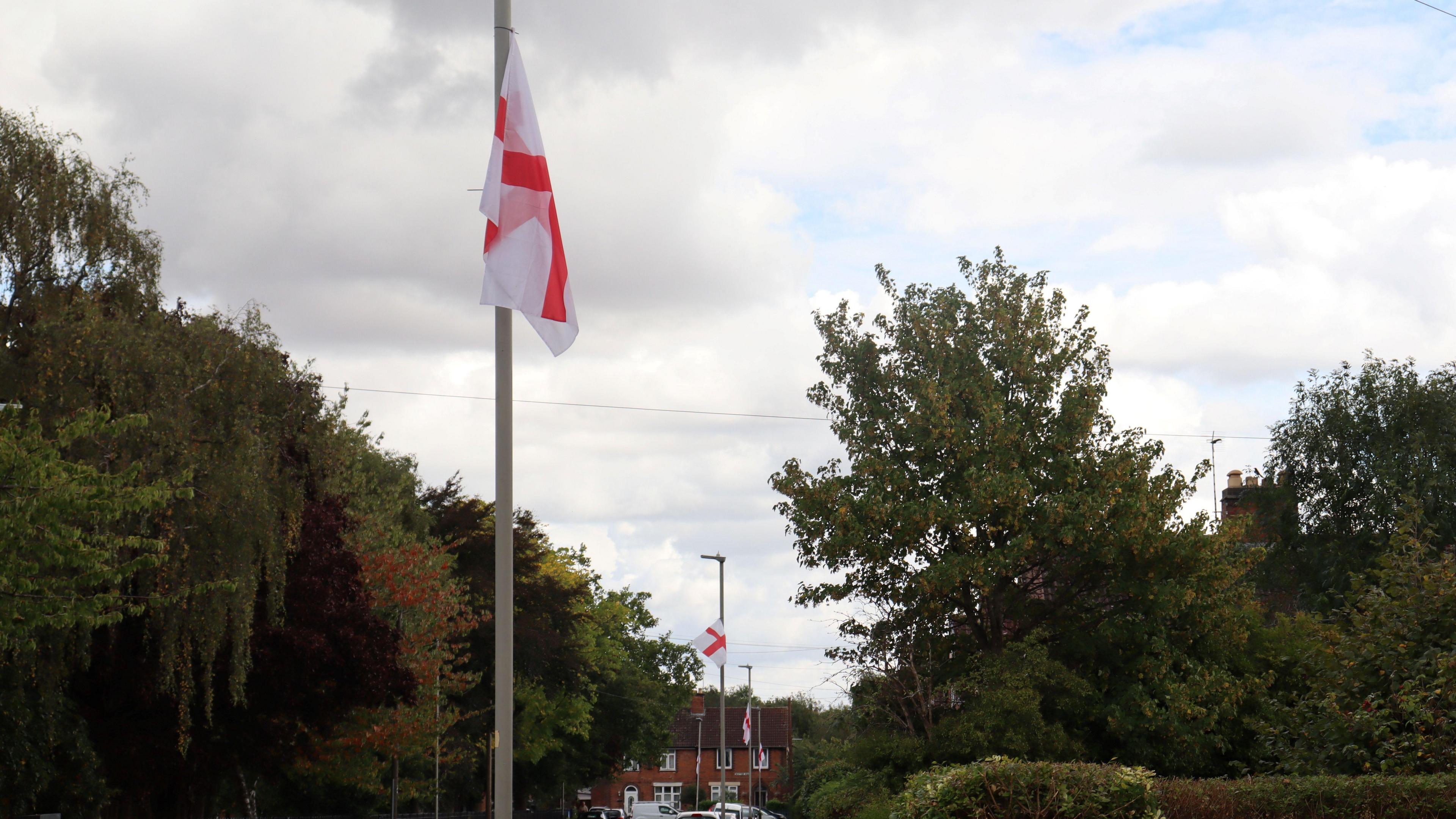 St George cross flags hung on a row of lamp-posts with trees on either side of the road.