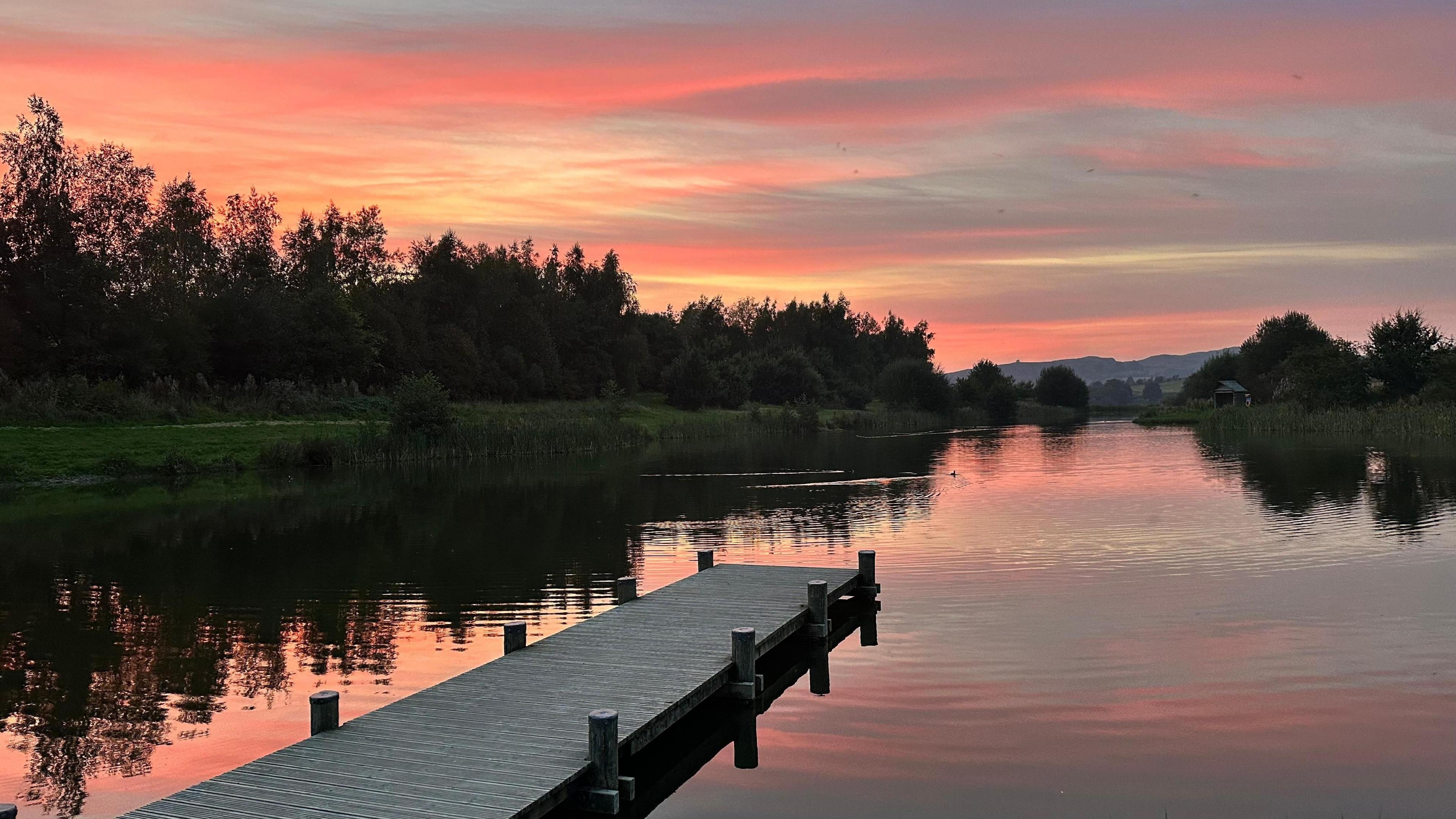 An orange sunset skyline above a body of water with a pontoon stretching out into the middle of the image. Trees can be seen lining the outside of the water.