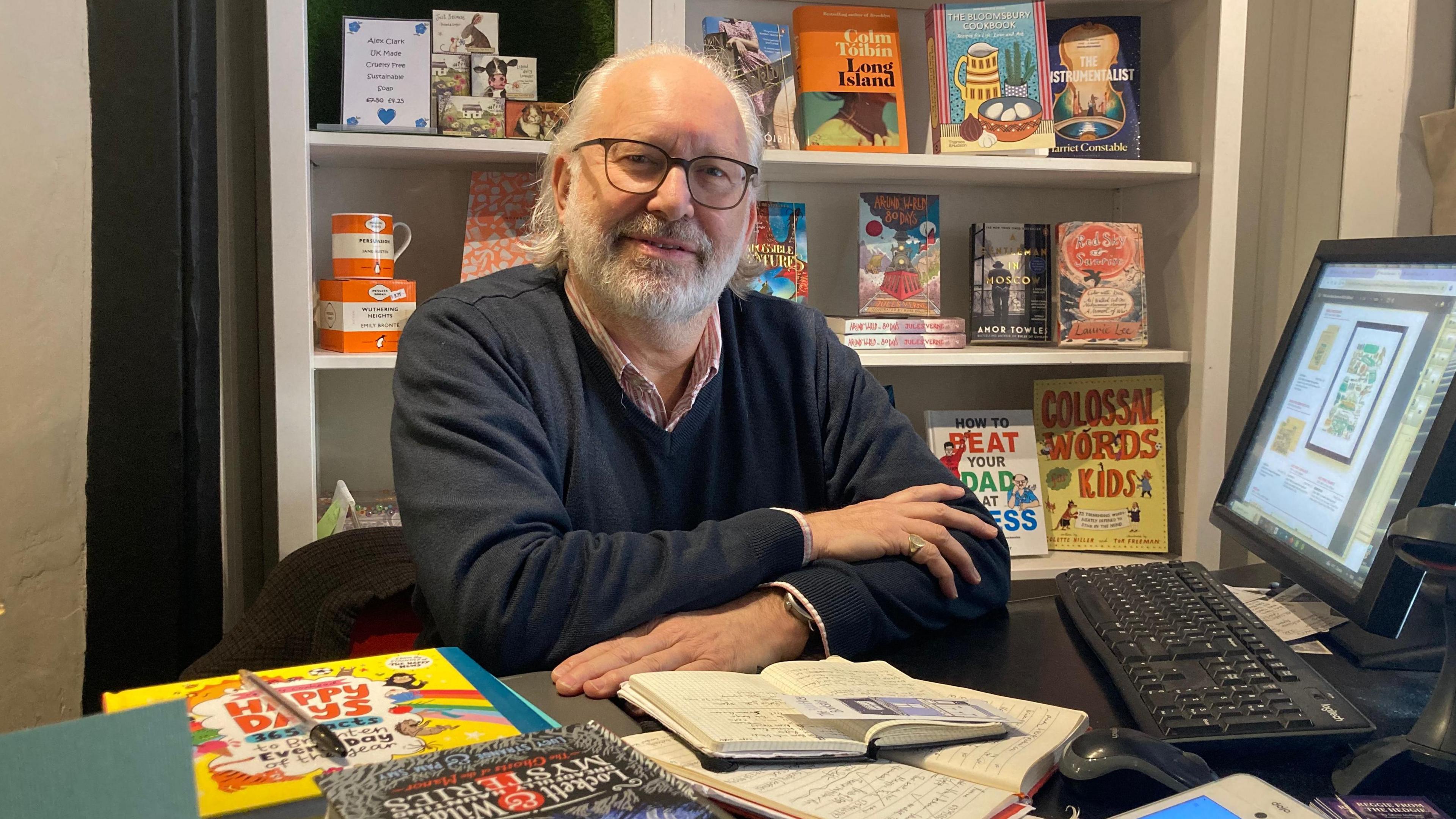 A bookshop owner sits at a desk, with books and a computer nearby. The man has a white beard and long white hair and is wearing black rimmed glasses. He has a pink shirt and a blue v-necked jumper on. 