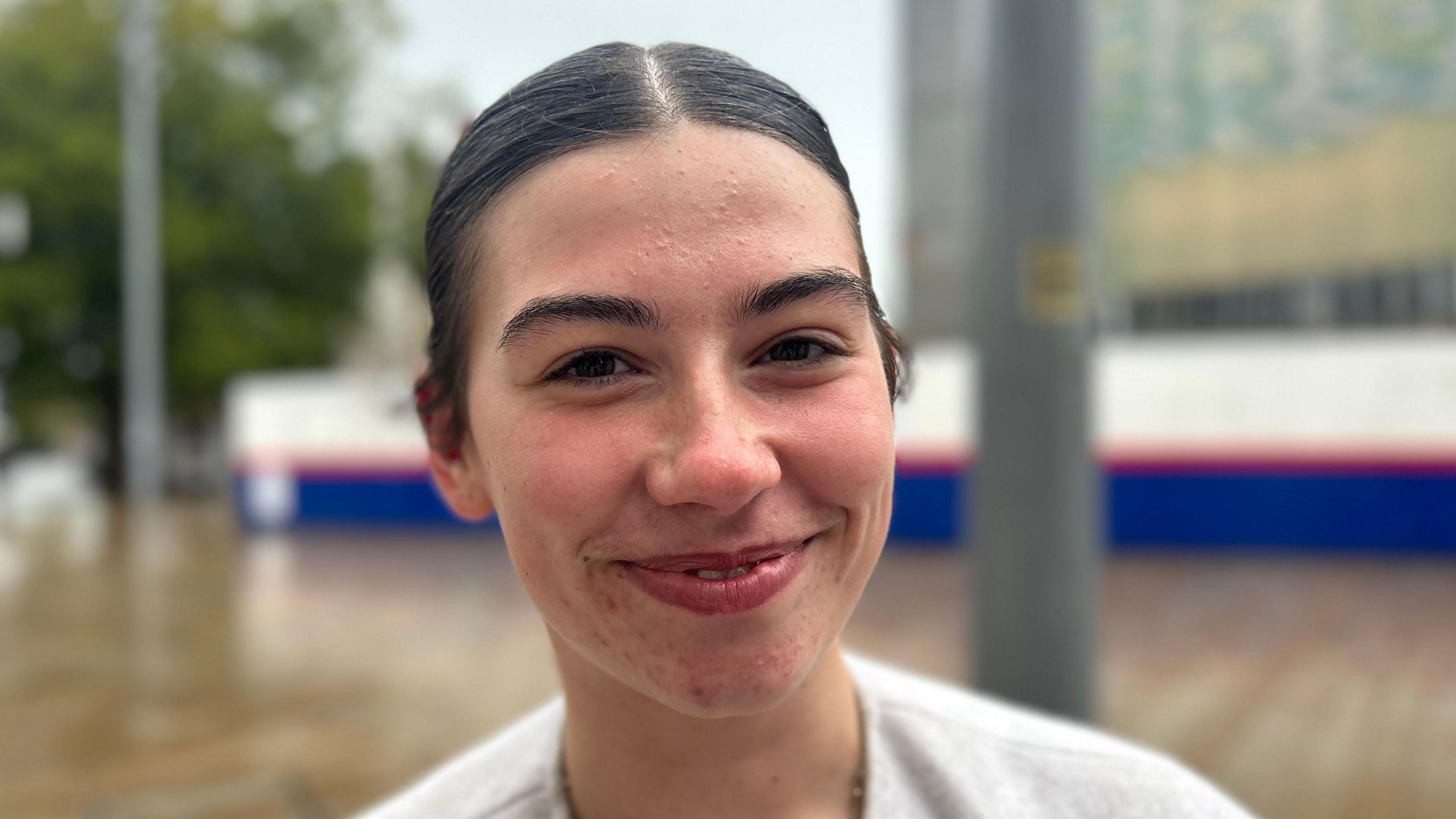 A girl with dark her and eyebrows, she's smiling in the middle of Hull city centre and her hair is slick back. It's in a pony tail and it matches her dark brown eyes. She looks in her early twenties. 