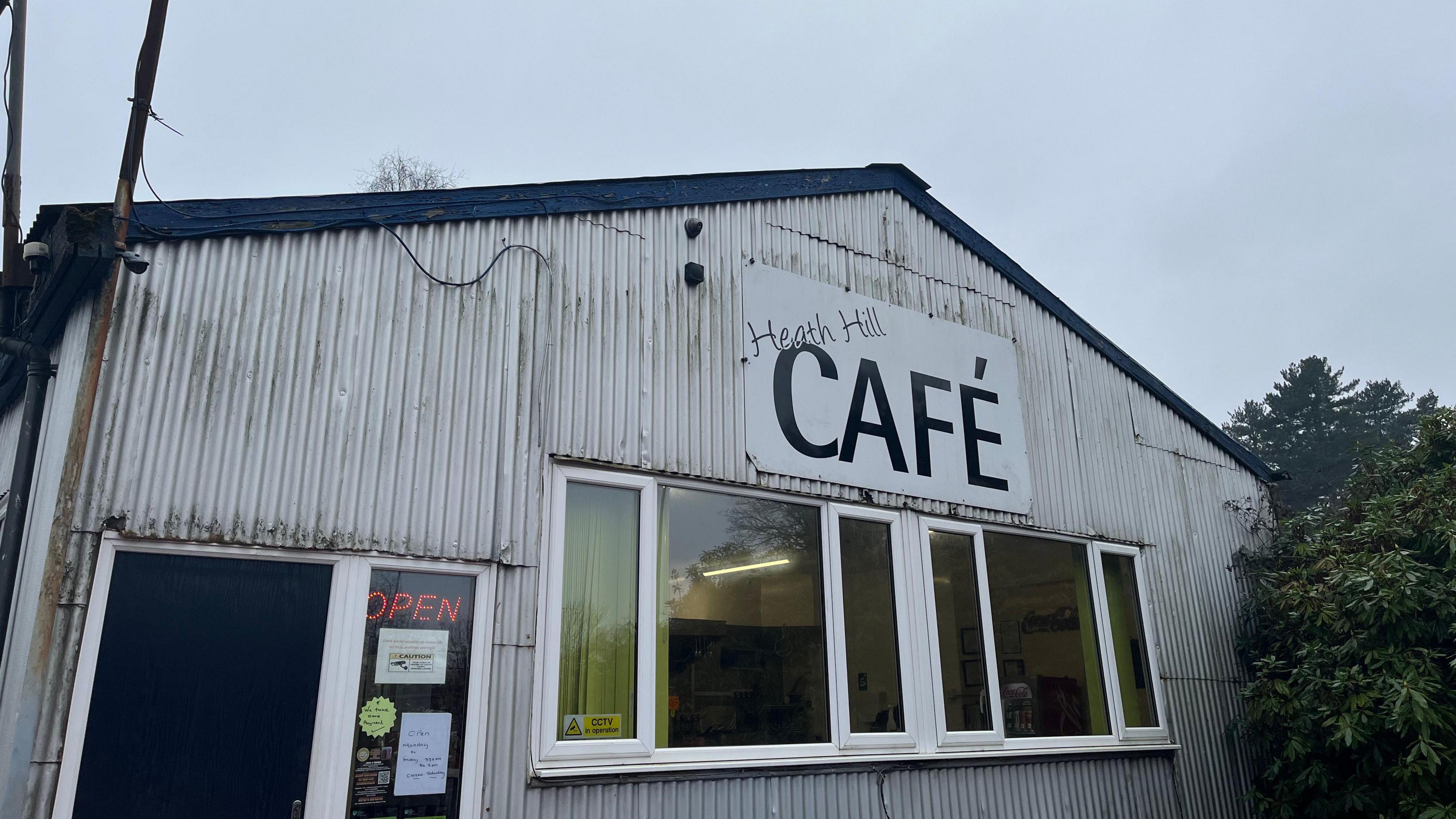 A small building clad in white corrugated iron with a black roof. It has a large sign on the front reading "Heath Hill Cafe" in black lettering. There are two large windows on the front where you can just see into the cafe, with chairs, tables, and a kitchen. On the left, there is a door with a neon sign reading 'Open.'