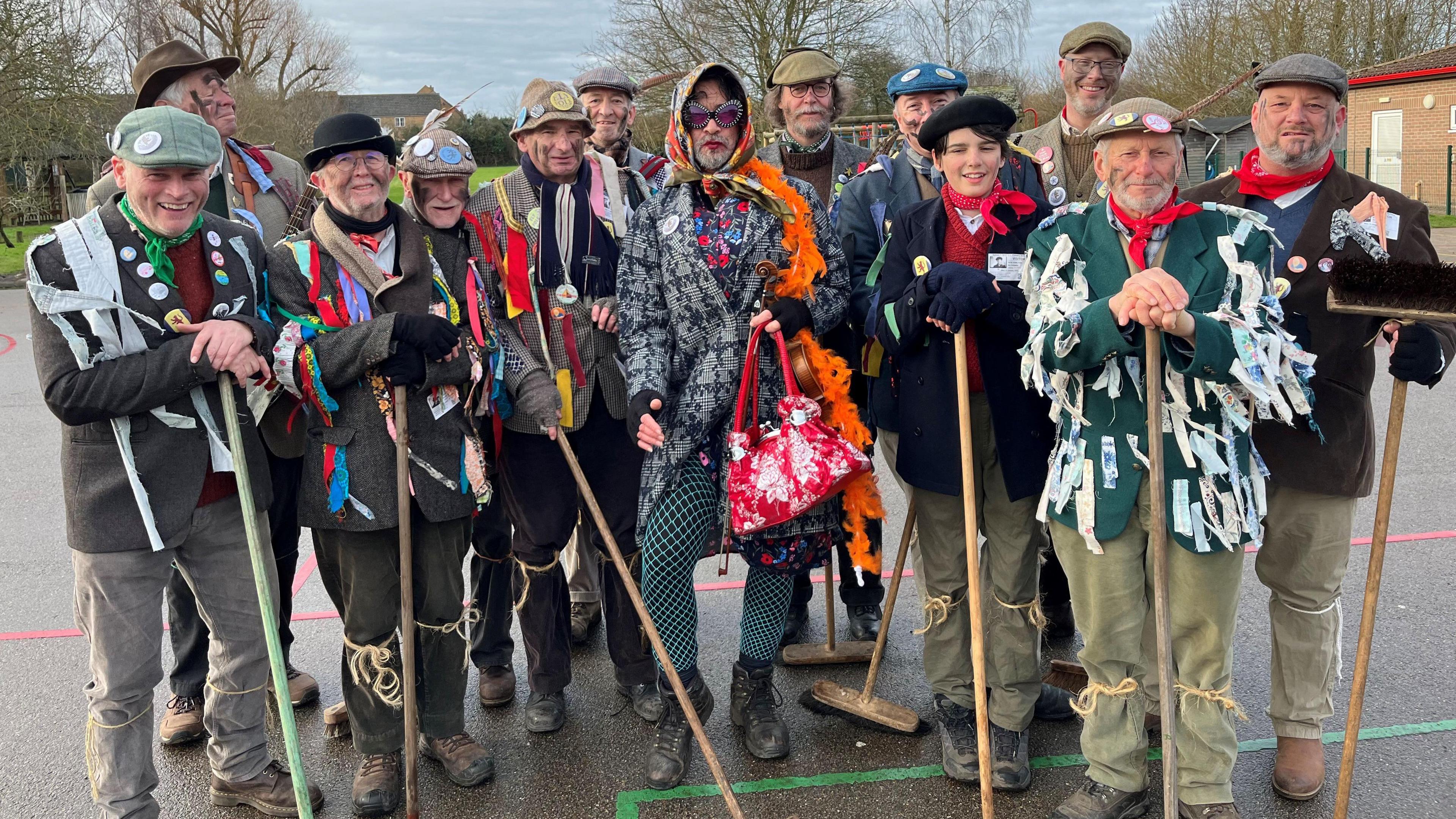 Members of Mepal Molly, a group of musicians and dancers standing in a school playground. They are all wearing traditional farming outfits that have been decorated with colourful ribbons, some are carrying brooms, and some have black soot smeared on their faces