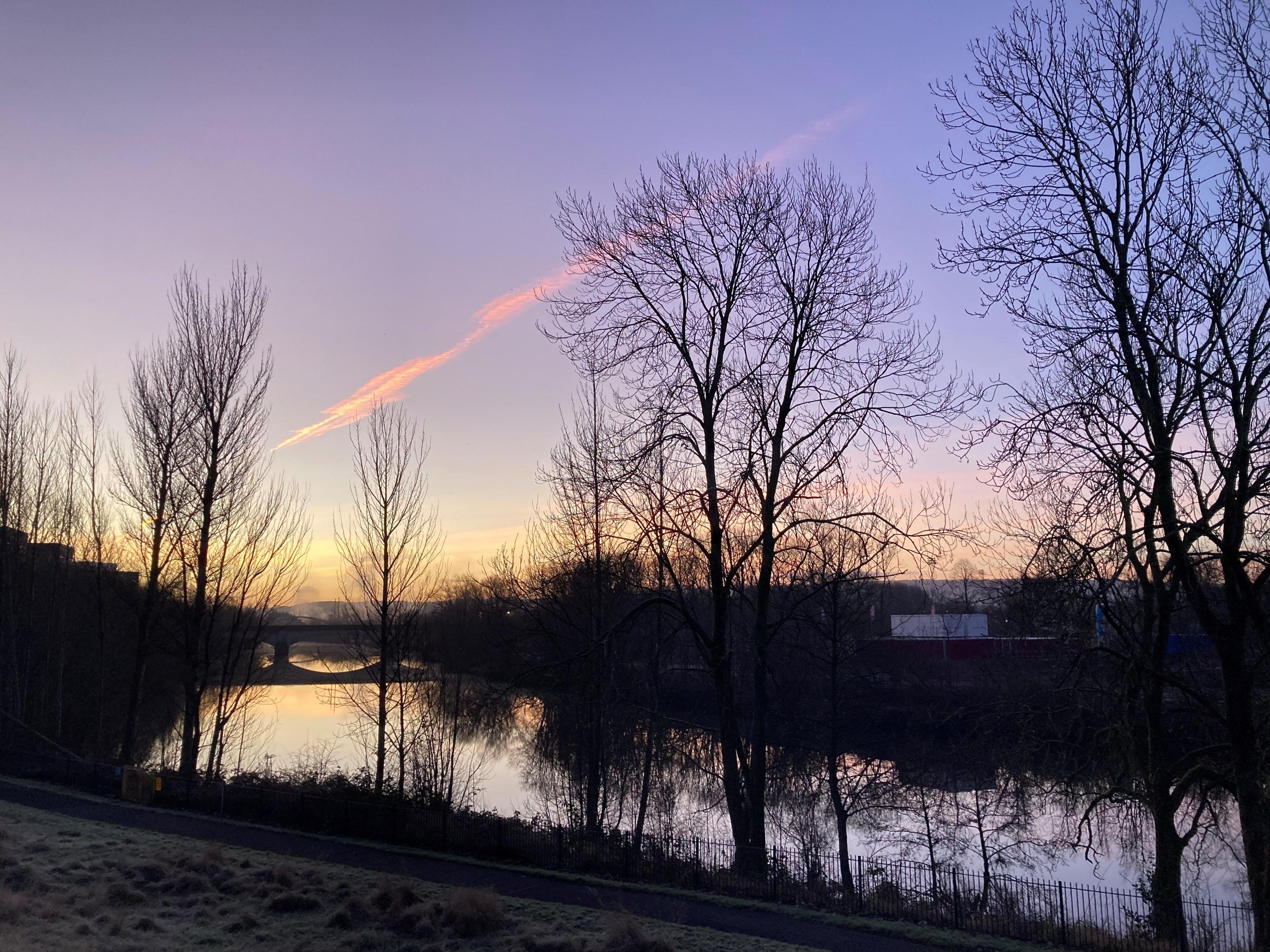 A wintry morning along the River Clyde, with a pink sky, sparse trees and grass with frost on the grass 