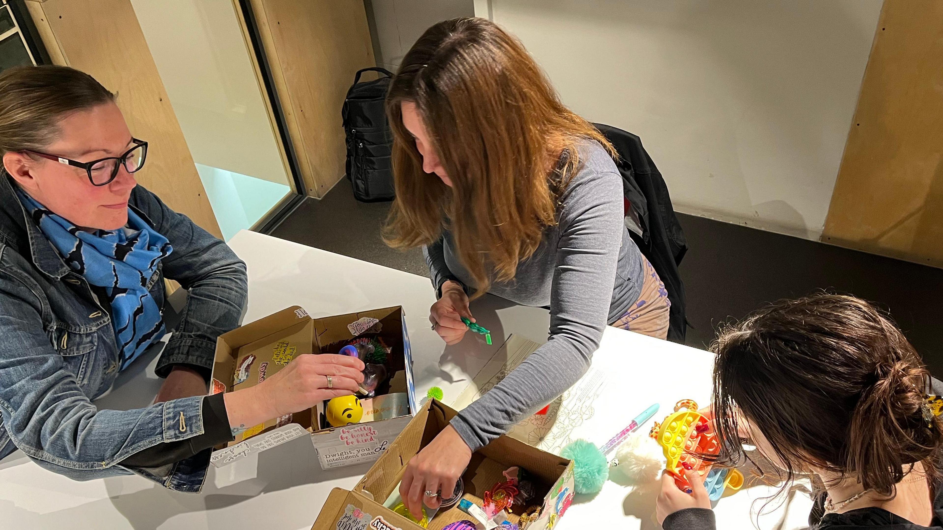 Two women and a teenage girl sit around a table taking fidget toys out of two open shoeboxes.