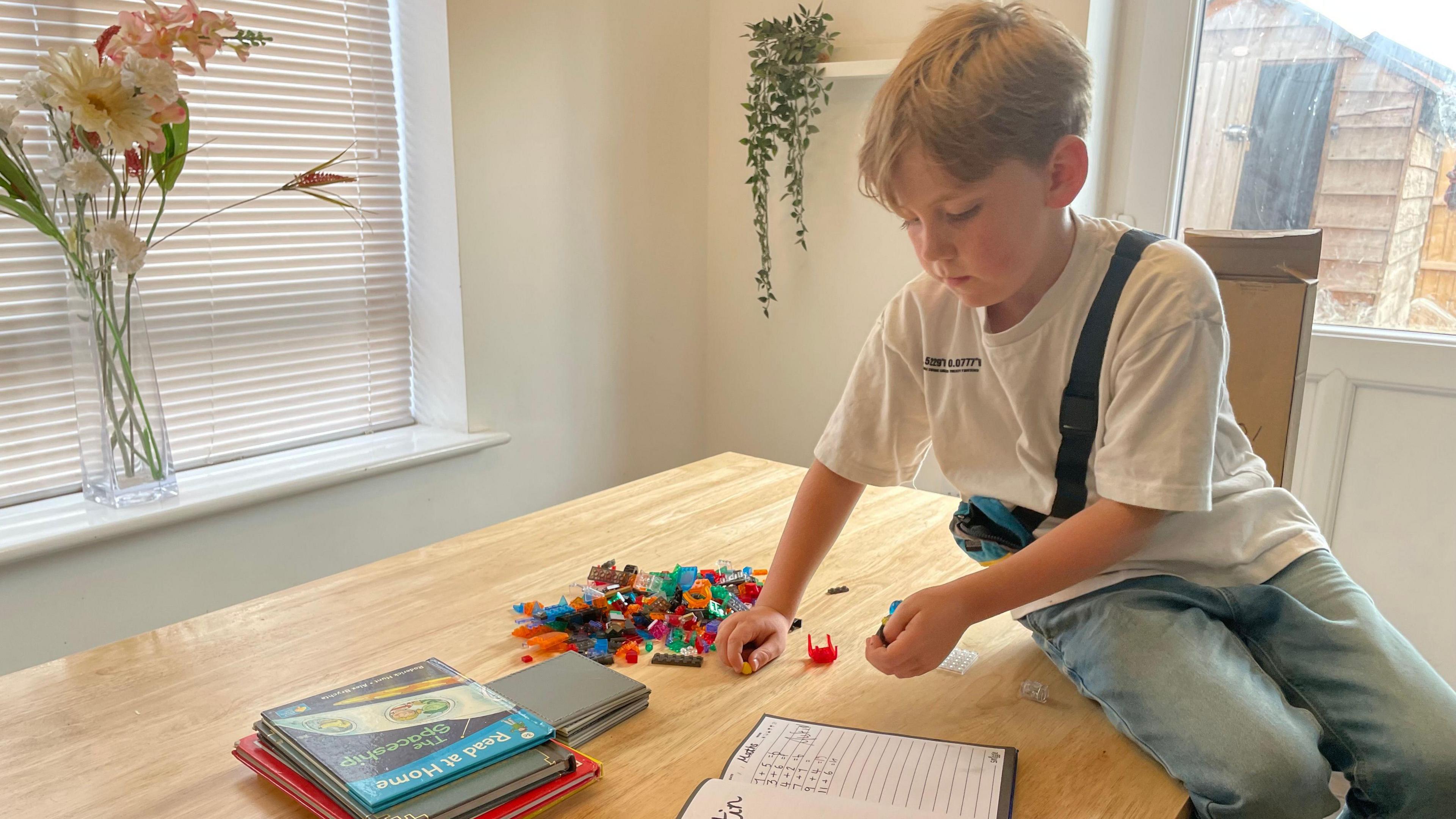 Austin sat on a wooden table which has books and LEGO blocks in a pile.