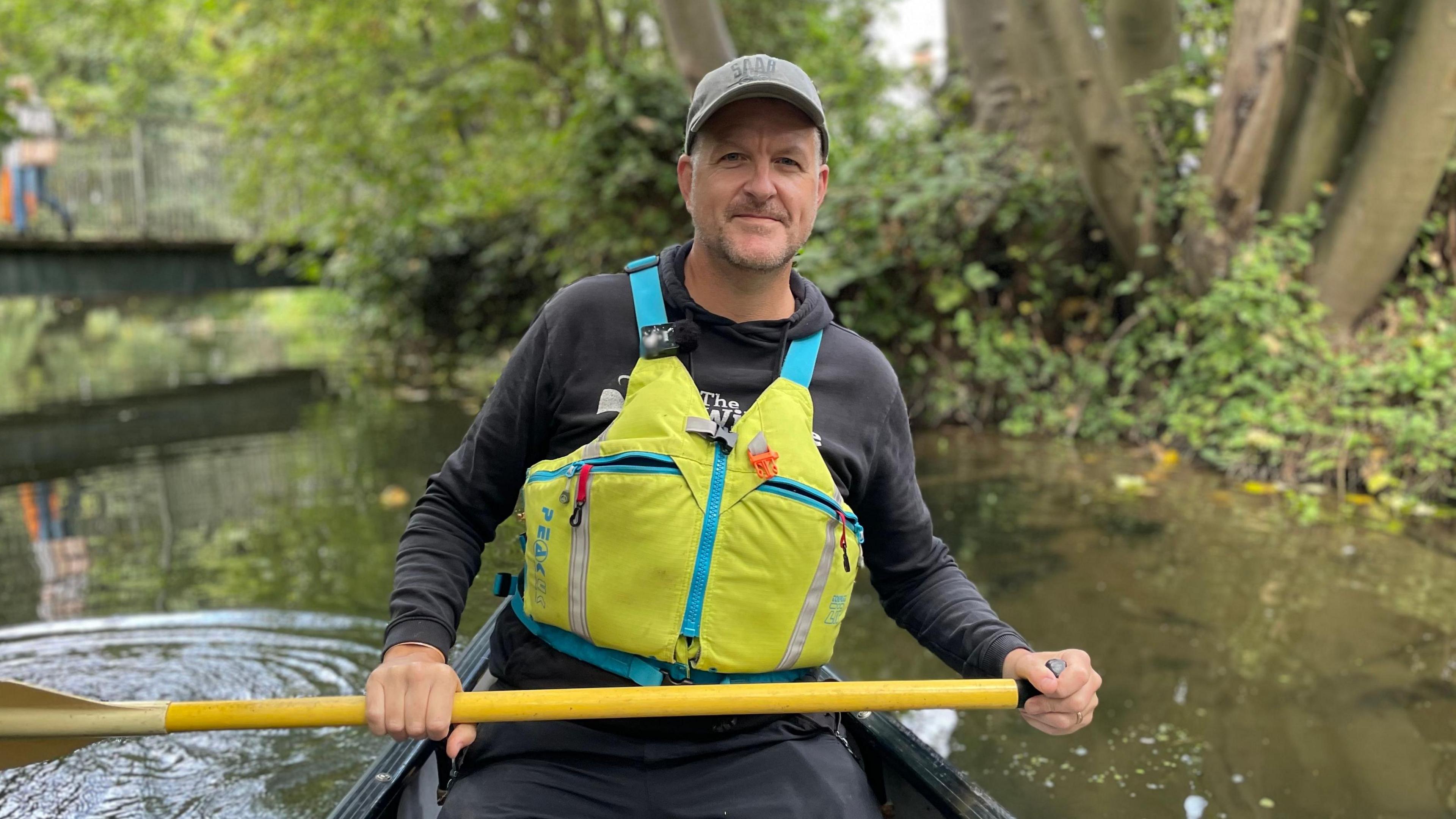 A man in a canoe, wearing a black hoodie and trousers with a yellow life vest is holding a yellow oar. He is also wearing a grey cap and is making his way down a river.