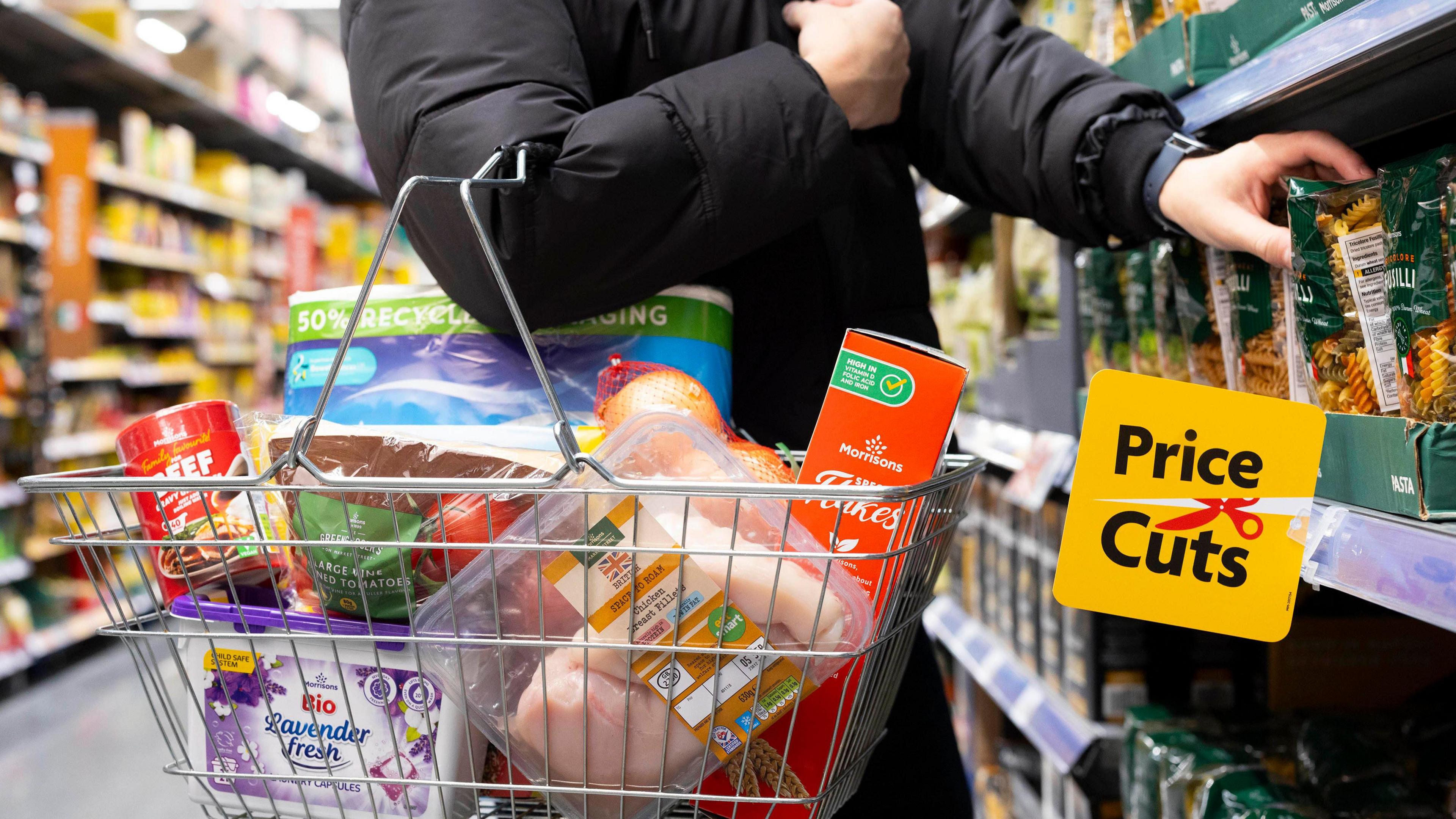 A shopping basket full of products being carried on a shopper's arm in a supermarket aisle. There is a sign on the shelf that says price Cut.