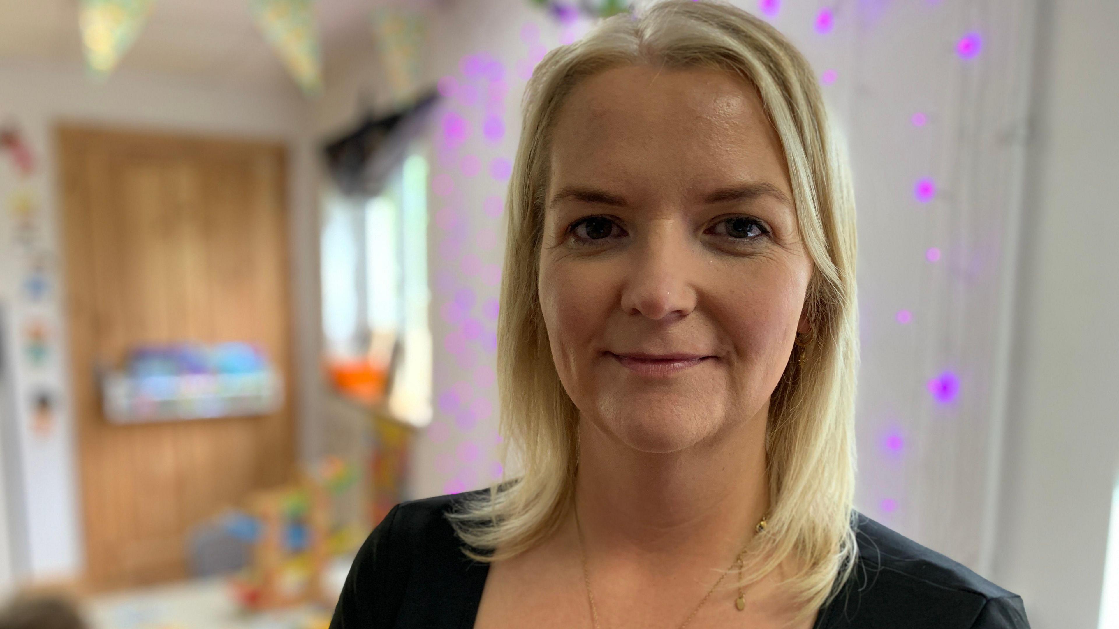 Theresa McCaughey has shoulder-length blonde hair and is looking at the camera. She has a gold necklace around her neck and is wearing a dark coloured top. There are children's toys in the background