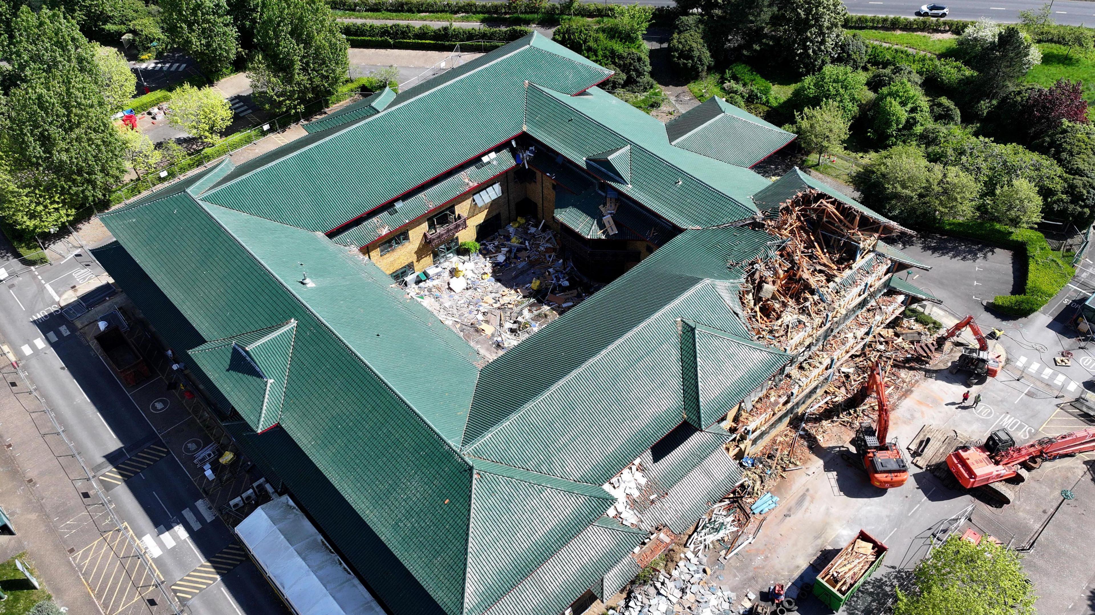 An aerial image of demolition works on a square building with a dark green roof. The building is surrounded by cranes and has a chunk taken out of its side. the roof has a number of triangular layers.