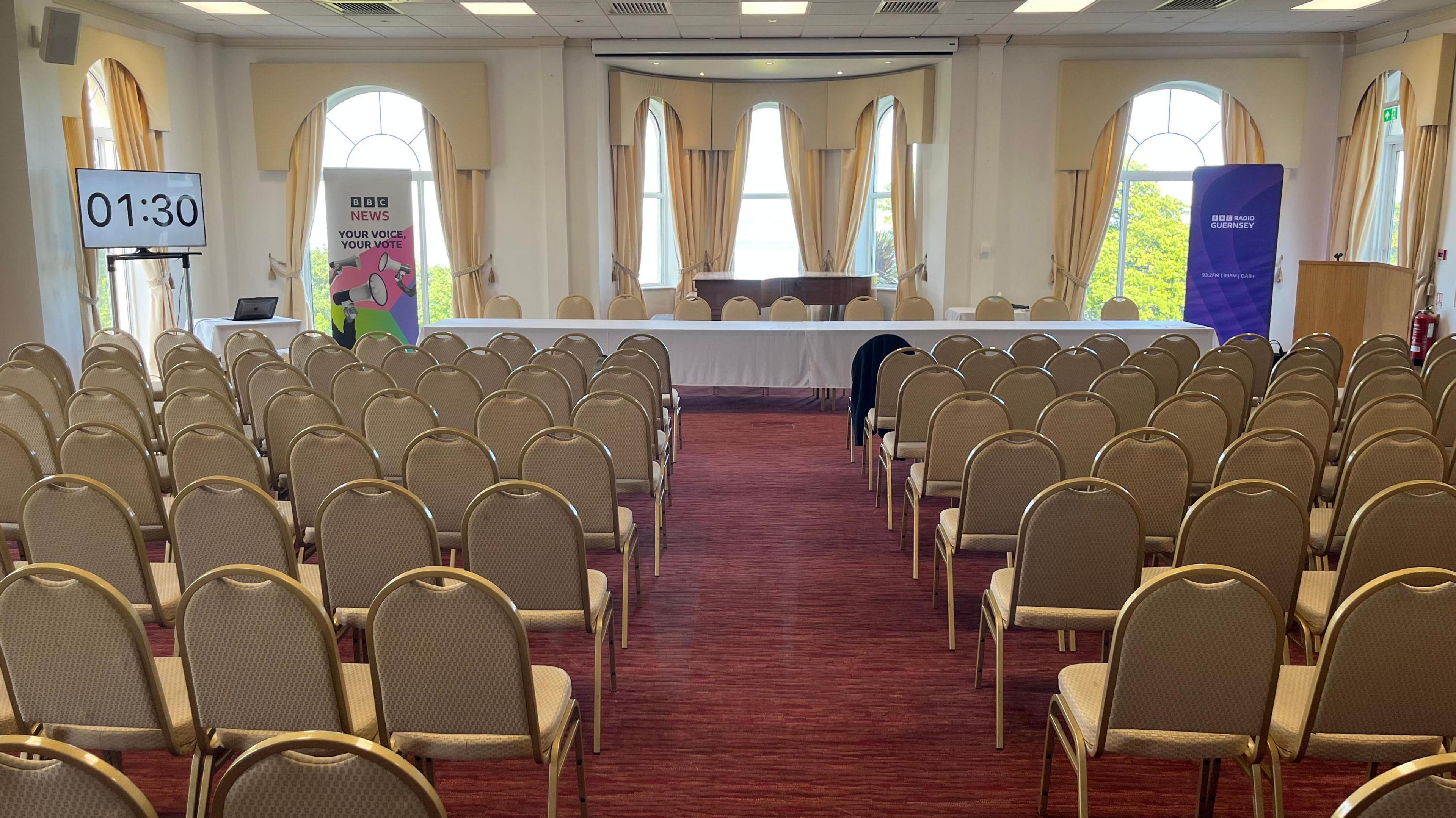 A room is full of rows of chairs and at the back is a purple banner which says BBC Guernsey and another that says BBC News Your Voice, Your Vote. On the left is a clock which says one minute thirty seconds. There are windows at the back of the room and pale yellow curtains.