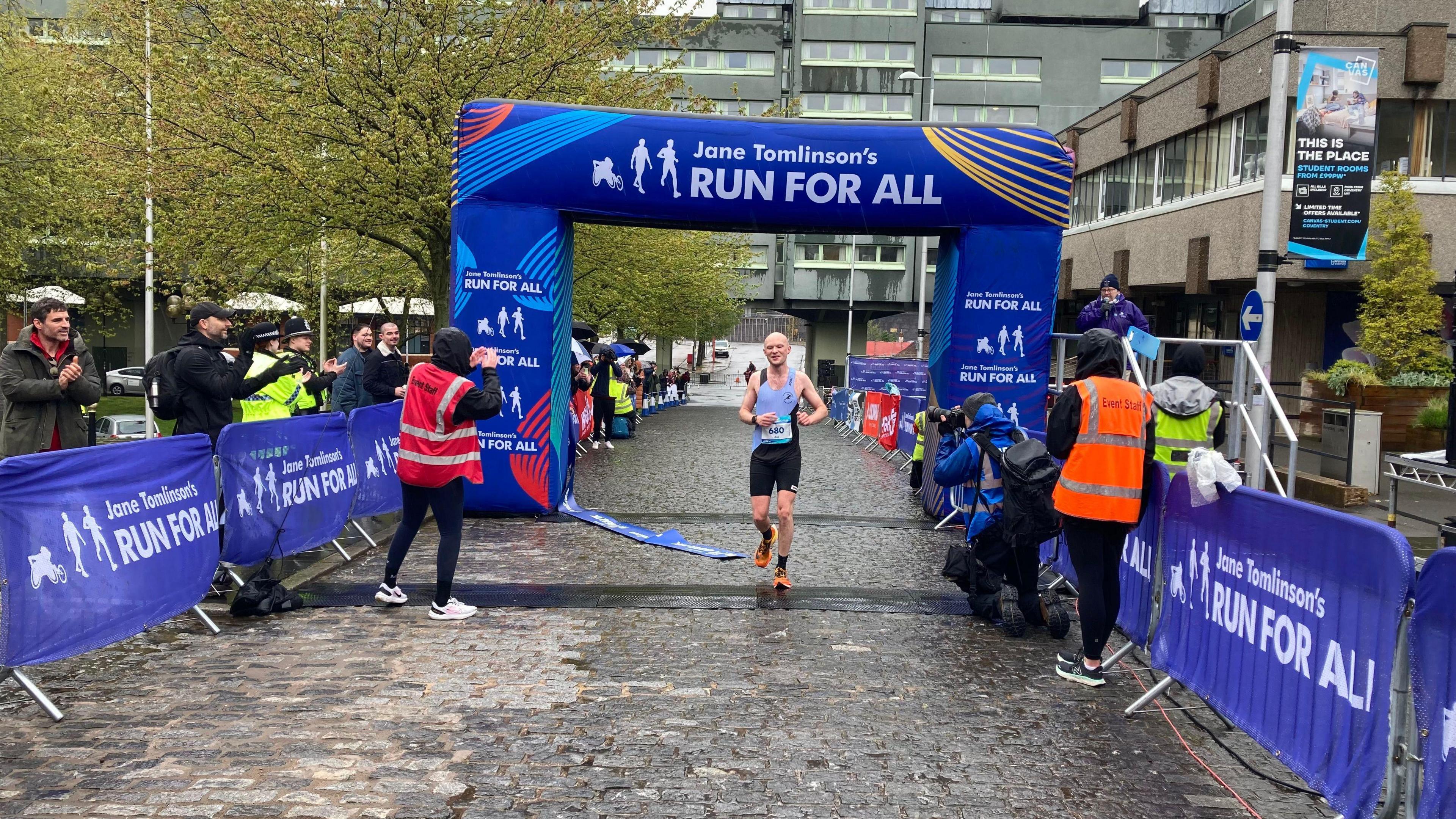 A man crosses underneath a blue finish line gantry in the Coventry half marathon. It is raining, and people are clapping 