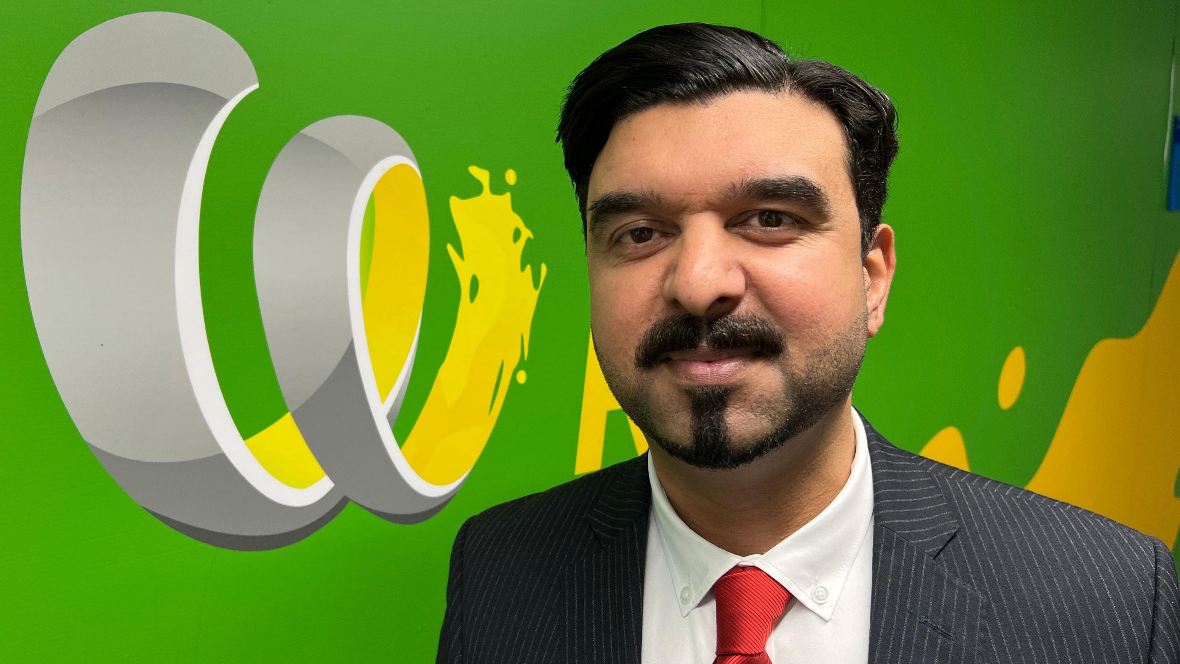 Councillor Kamran Caan is pictured in front of a silver, green and yellow logo for The Wave. He has black hair and beard and is wearing a red tie, white shite and grey pinstriped suit.