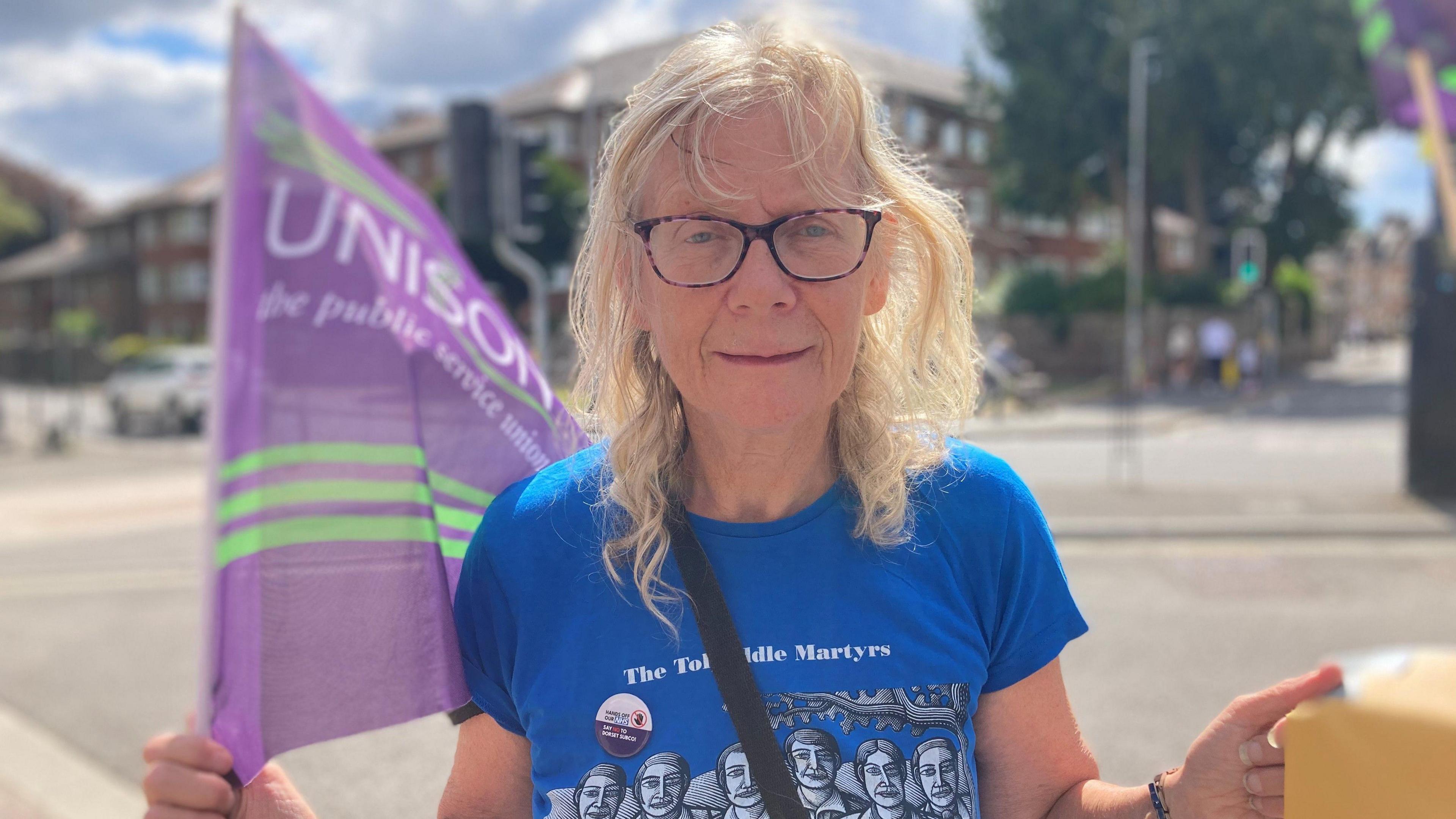 A head-and-shoulders photo of Lynne Hubbard, a woman with blonde hair, glasses and wearing a blue T-shirt. She's holding a Unison flag, and a road and building can be seen behind her.
