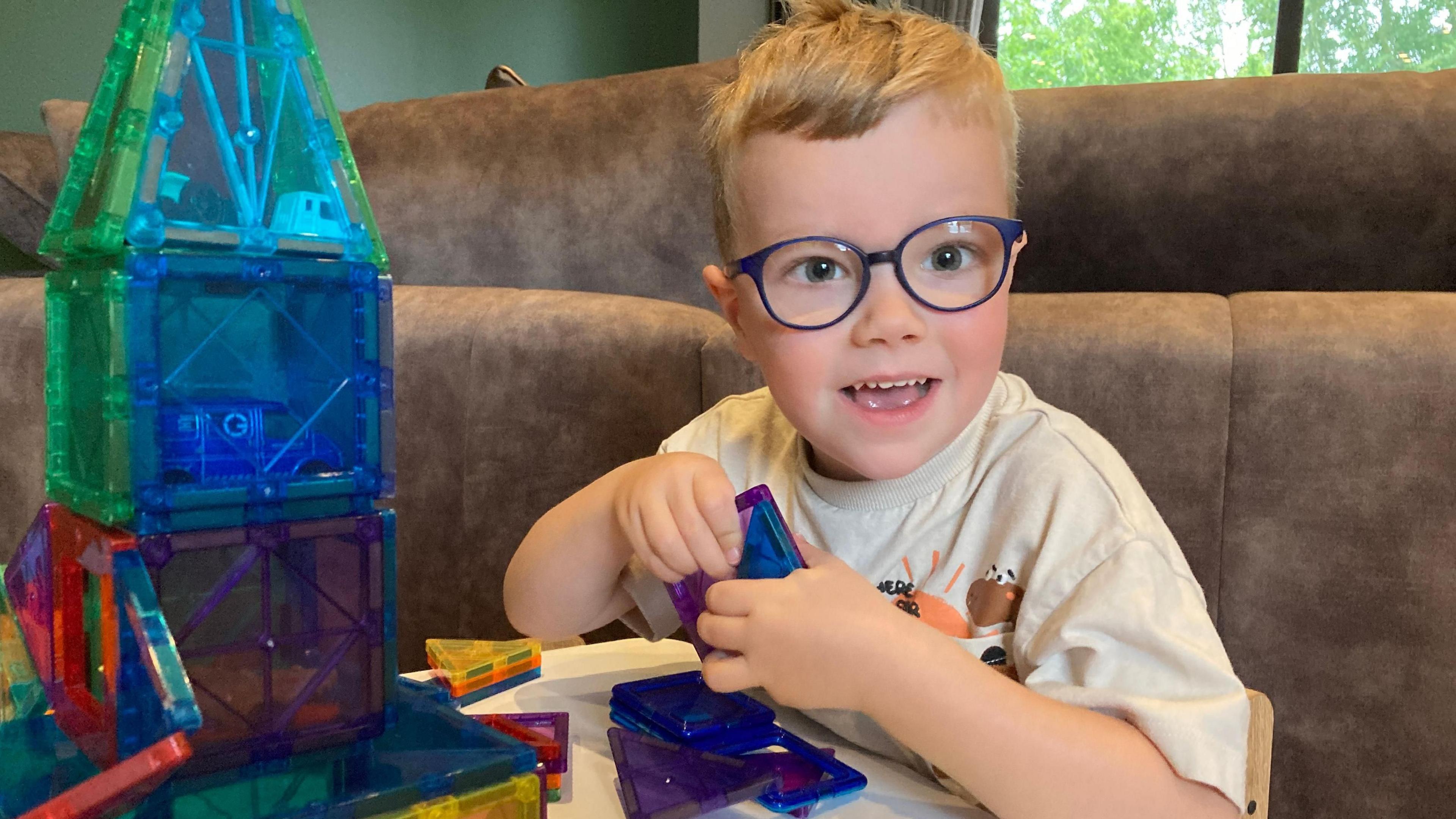 Max is smiling and wearing glasses. He is playing with a construction set at home and has made a tower.