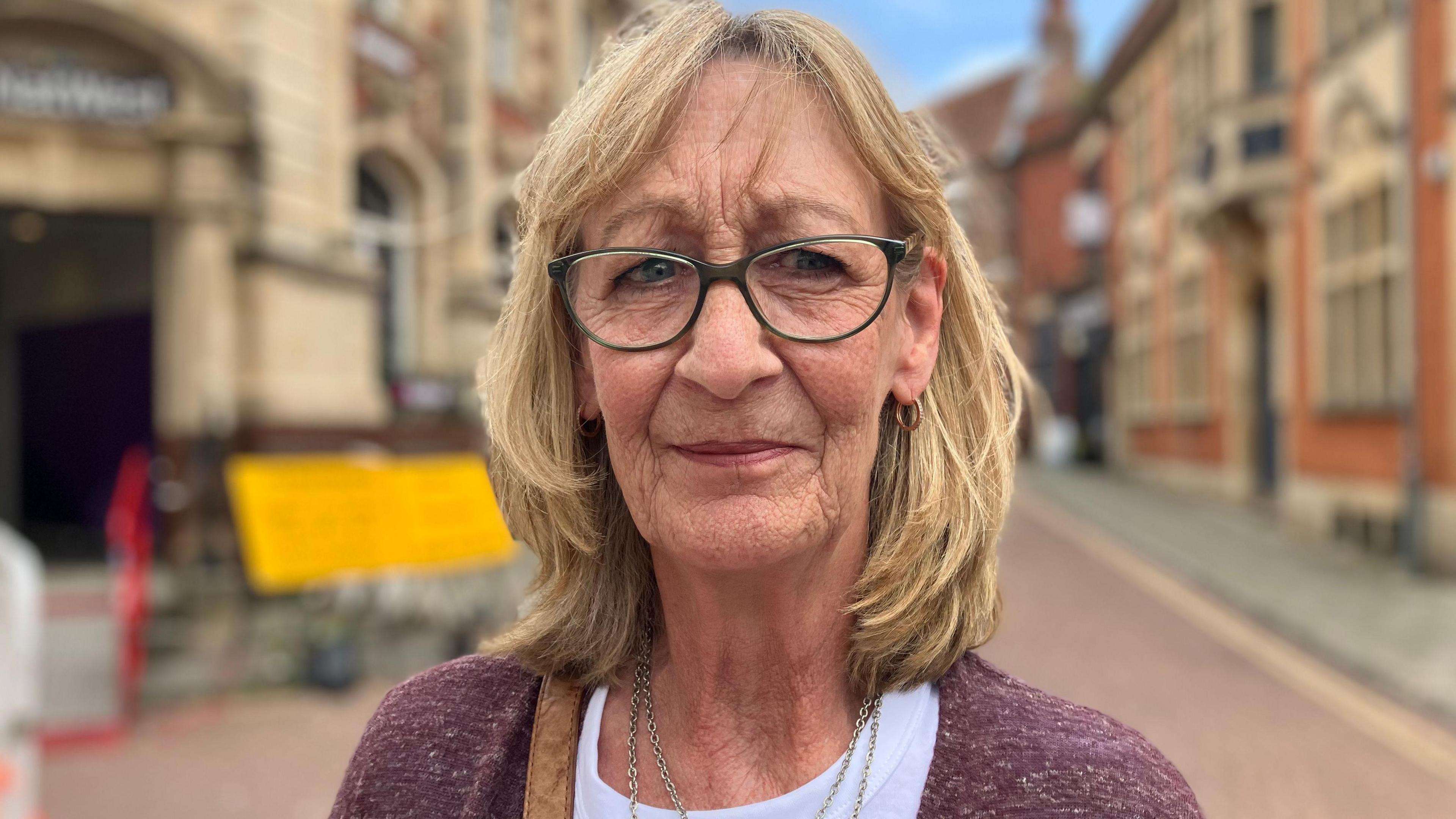Headshot image of a woman with blond shoulder length hair, wearing glasses and a purple cardigan standing on a street