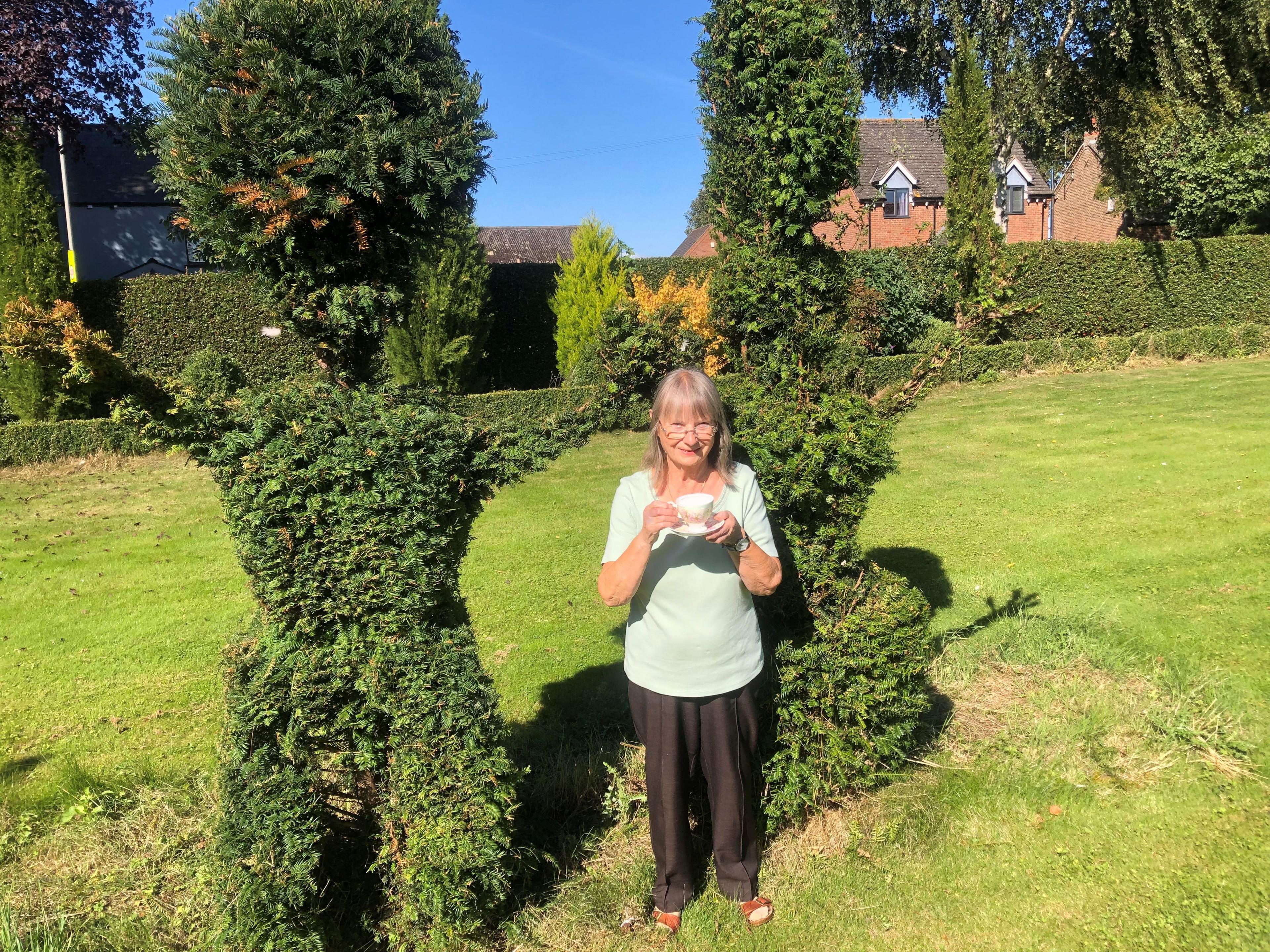 Ellen Beeton with a cup of tea stood in the middle of the topiary sculpture of herself and her late husband. She has long grey hair and is wearing a light blue T-shirt, black trousers and brown sandals.