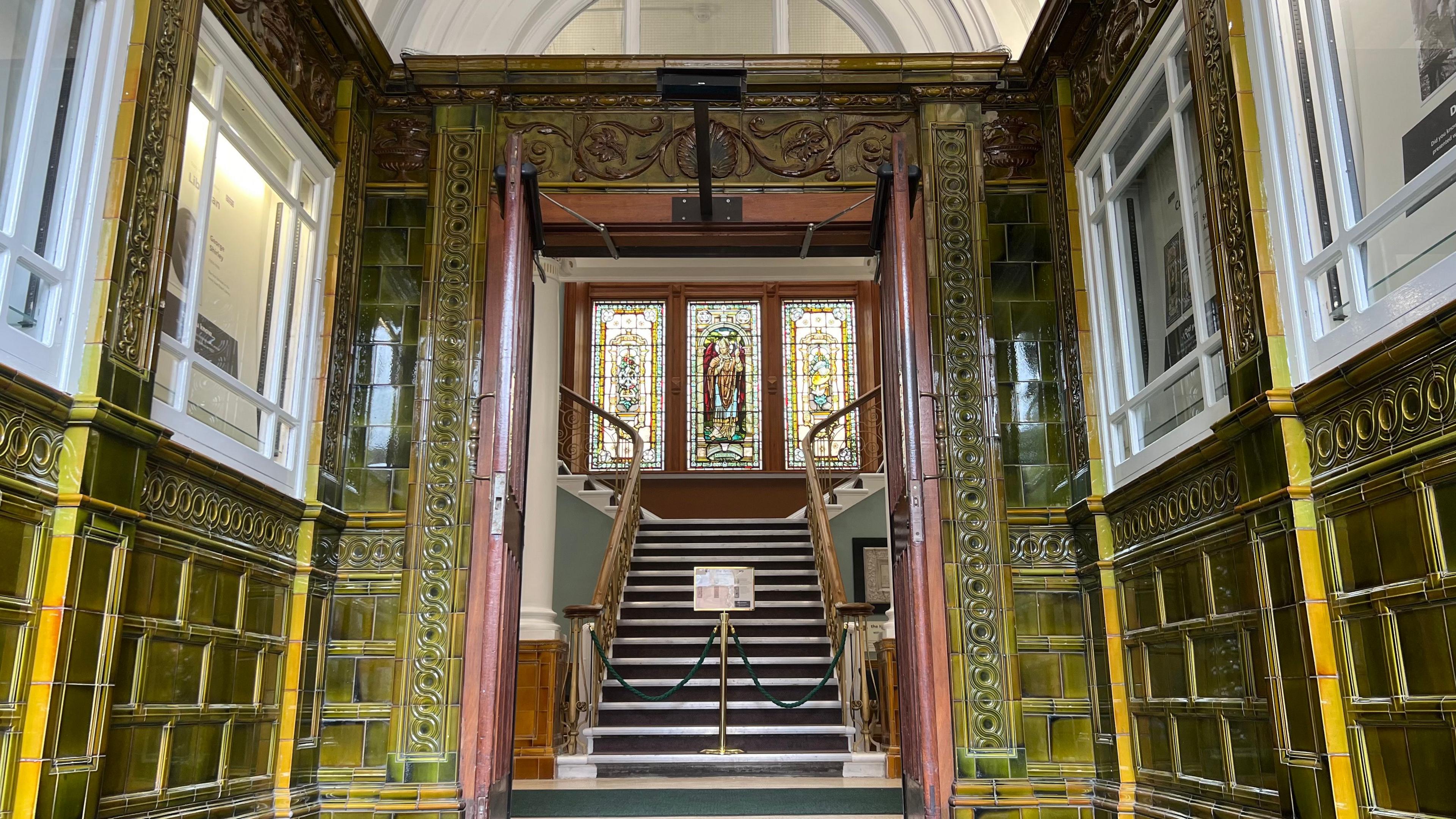 The entrance corridor of the library - it's covered in emerald-green tiles with a yellow reflection in the light. Looking through open dark wooden doors towards three bright and colourful stained glass windows above a staircase.