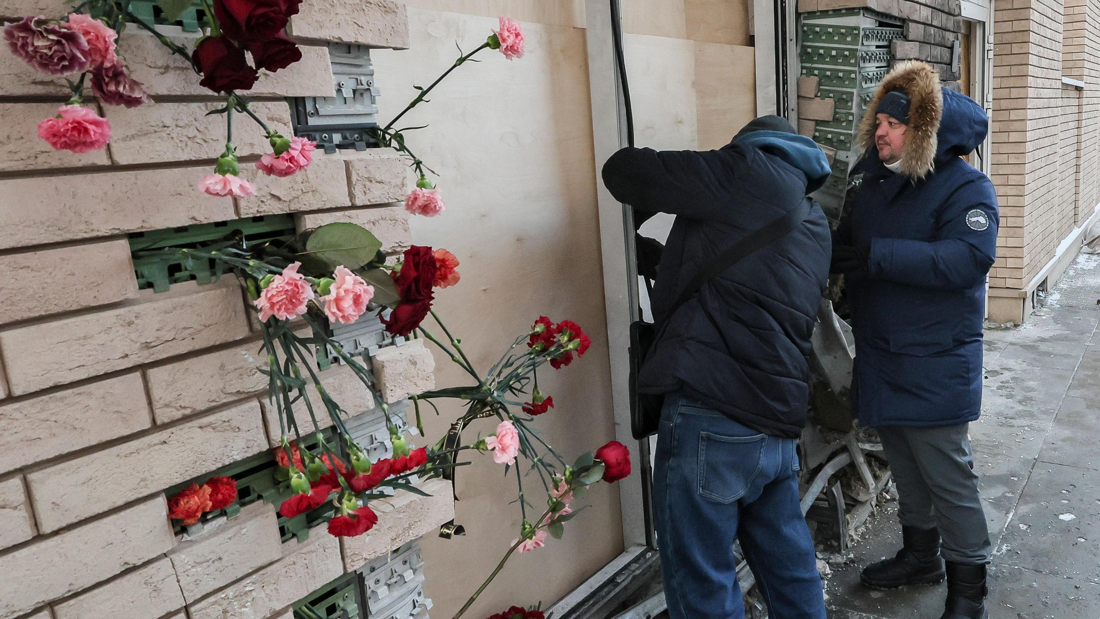 Municipal workers in puffer jackets inspect a blown-out door that has been boarded up outside the apartment building where Kirillov died. Plastic flowers are placed in exposed brickwork
