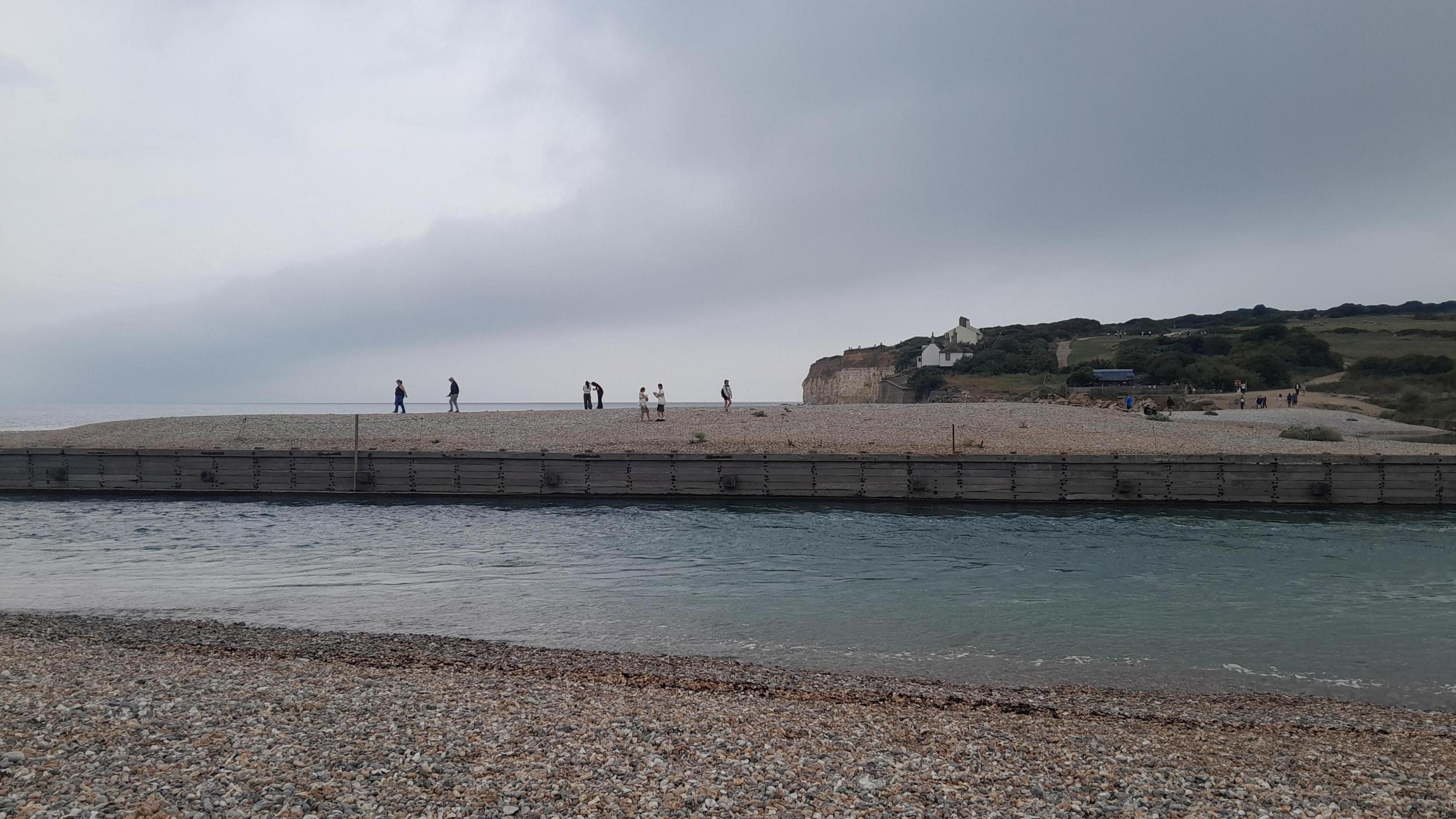 Seven people are pictured in the distance across the mouth of the River Cuckmere, with a shingle beach in the foreground.