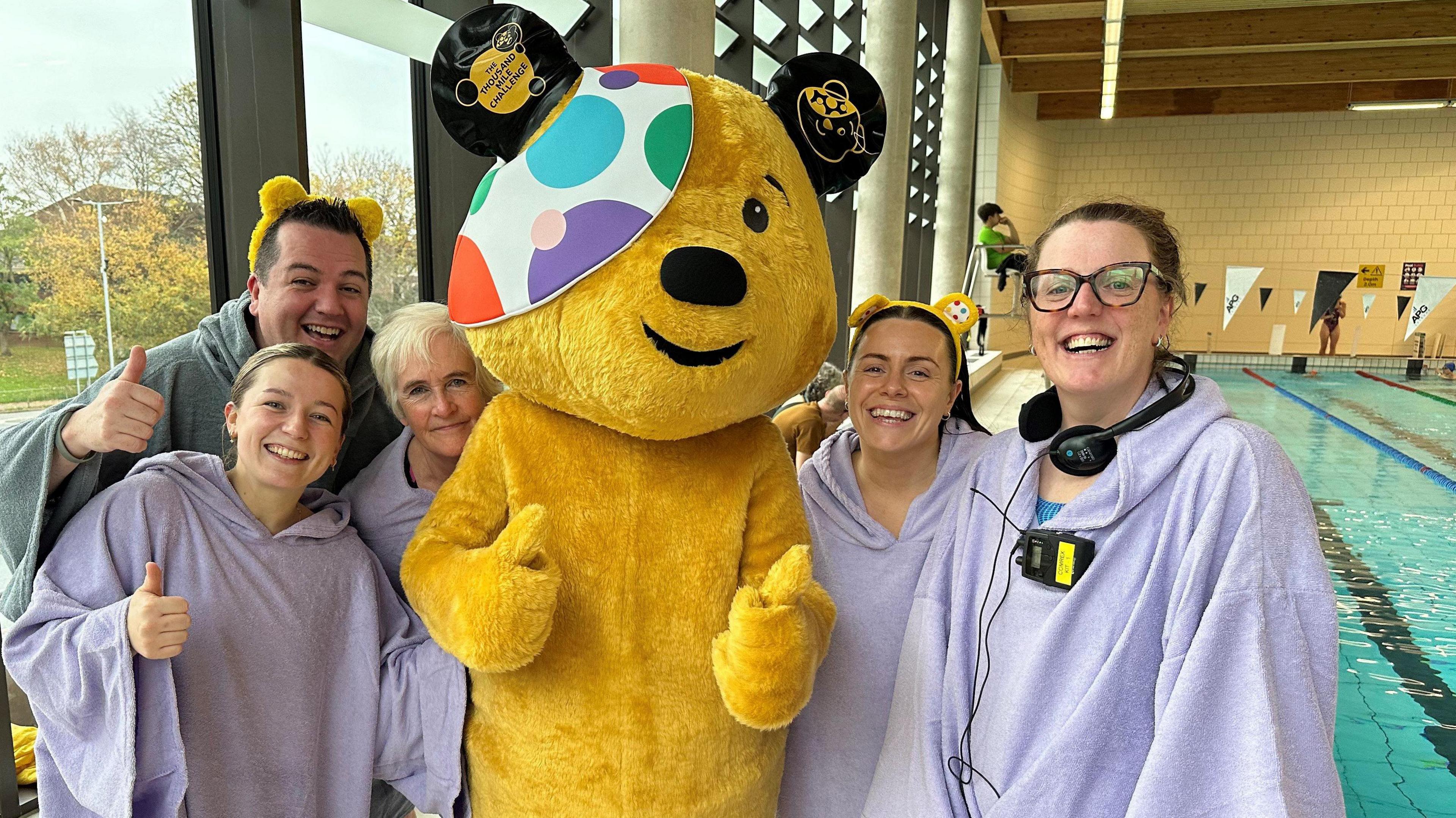 The five swimming team members in purple robes stood around Pudsey bear. Pudsey and some of the team are giving the thumbs up. The pool is in the background. 