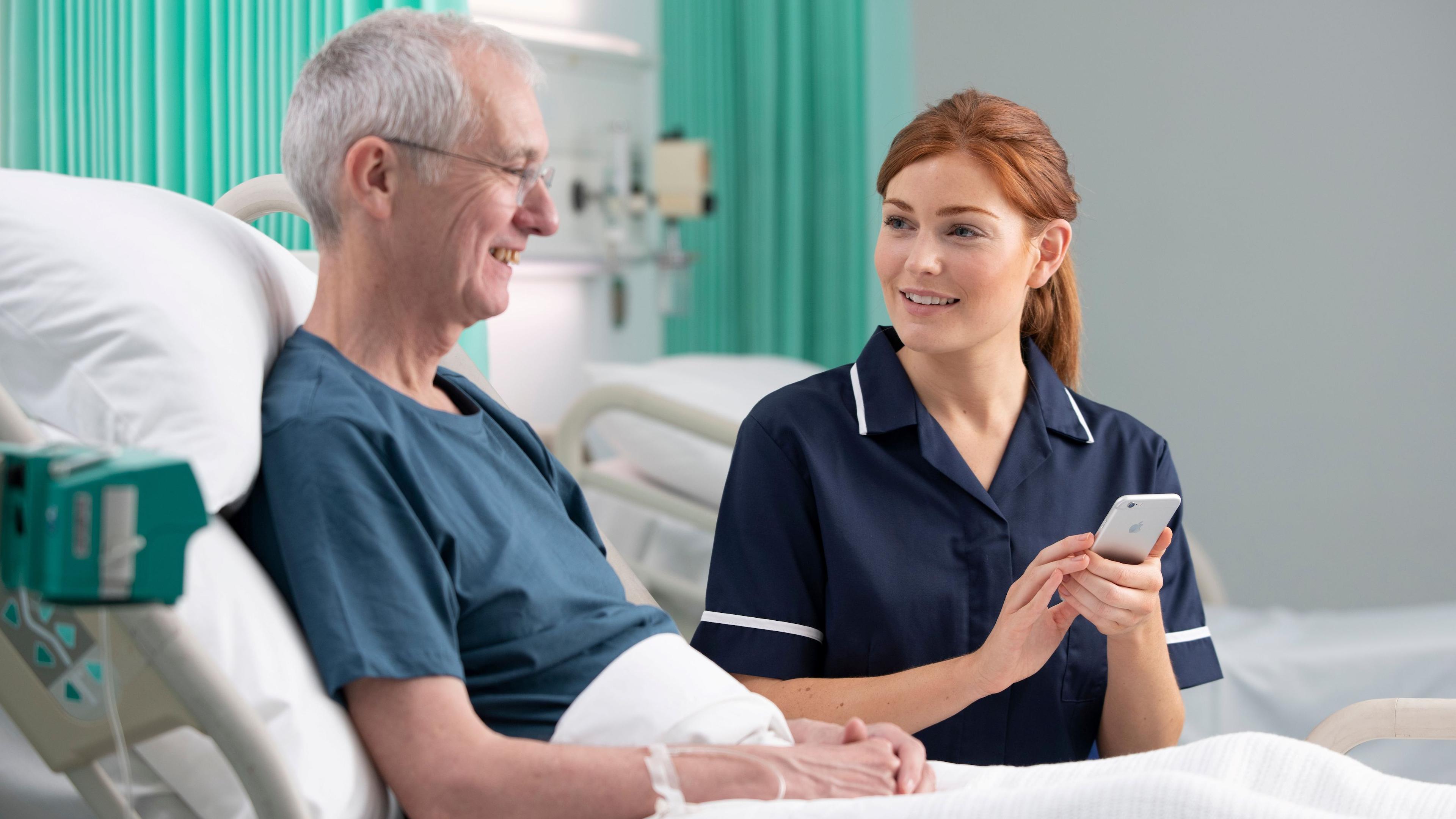 A man, in a hospital bed, wearing a blue top, with short grey hair and glasses, smiling, with a nurse, in a blue uniform, with long red hair, tied back, smiling at him, while holding a phone. There is a bed behind them, all with white sheets on.