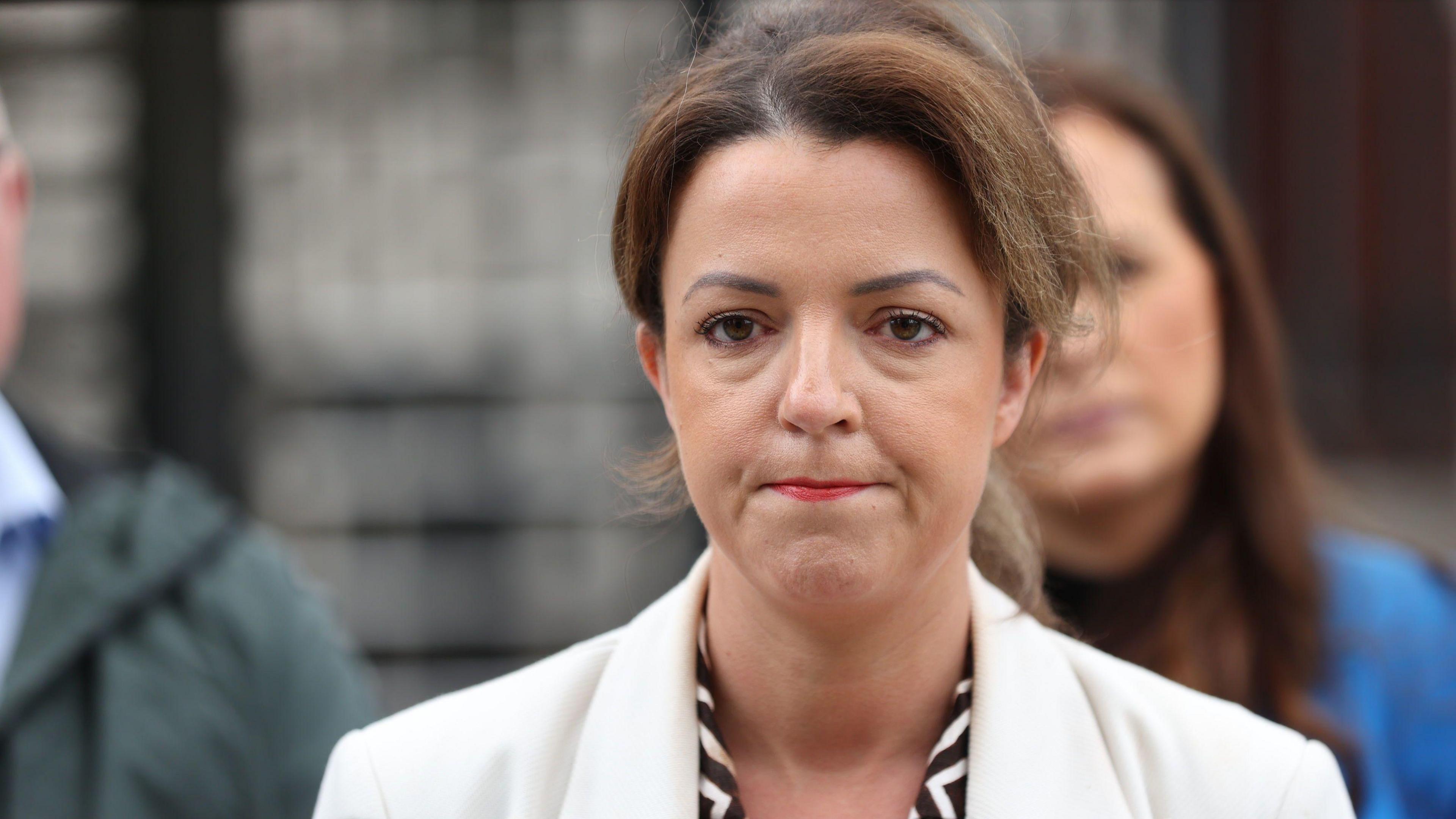 A close-up image of Liz Kimmins standing outside court. She has her lips pursed together while looking at photographers. She has dark, highlighted hair which is in an updo, and she is wearing a white blazer and a black and white patterned shirt.