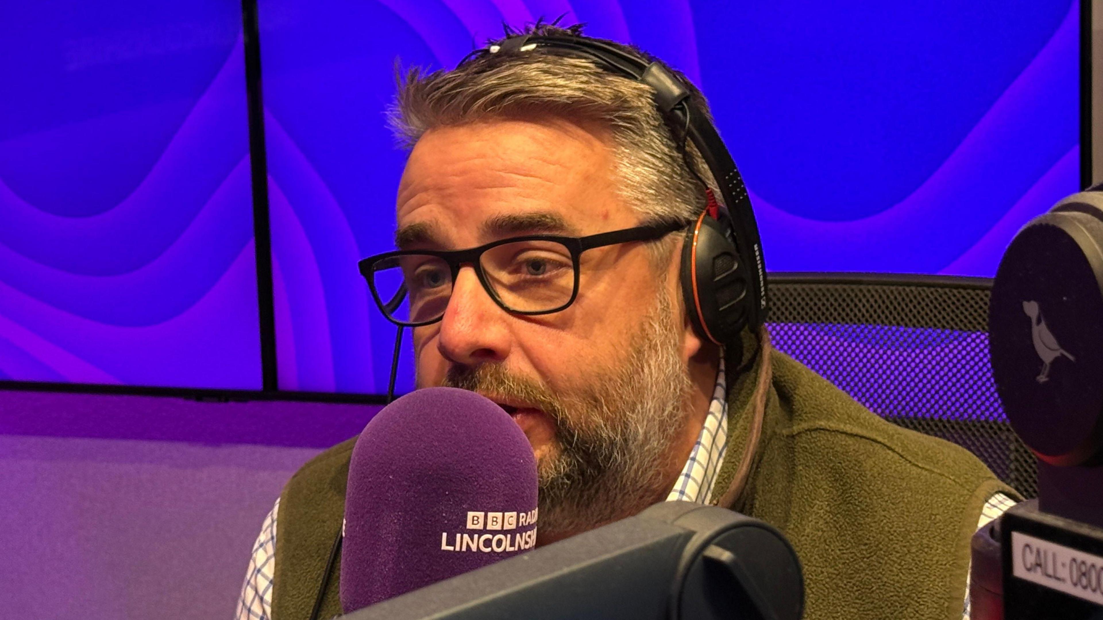 Richard Davies has grey hair and a grey and brown beard. He's wearing a green jacket and is sitting in front of a purple microphone. Behind him are an array of purple screens.