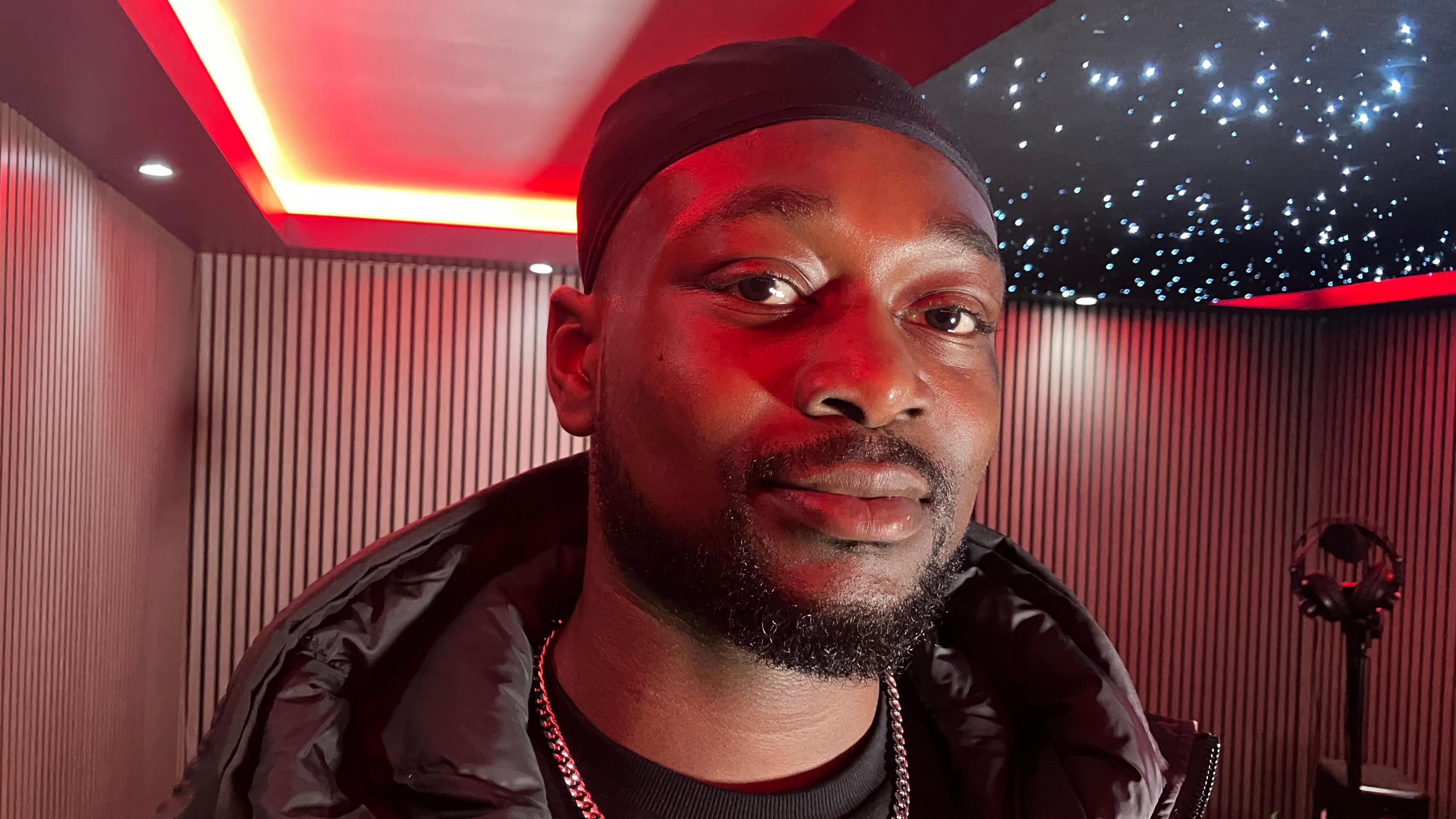 FSJ. He has a beard and dark eyes, wearing a black bandana, black coat and goad chain. He is standing in a recording studio with red lighting.