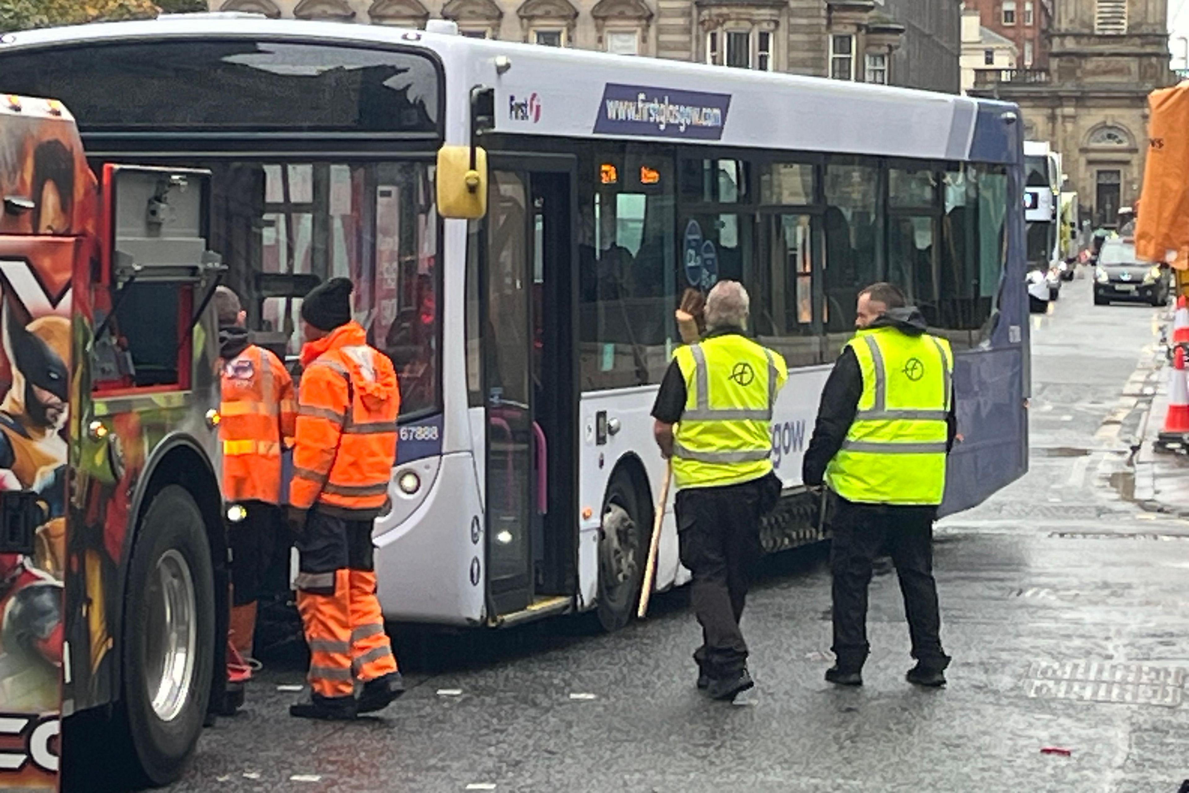 A First Glasgow bus that has crashed into a building in George Square. The front of the bus is damaged, and workers in high-visibility orange and yellow clothing are gathered around it. Another vehicle with superhero artwork is positioned in front, assisting at the scene. Historic stone buildings surround the square.