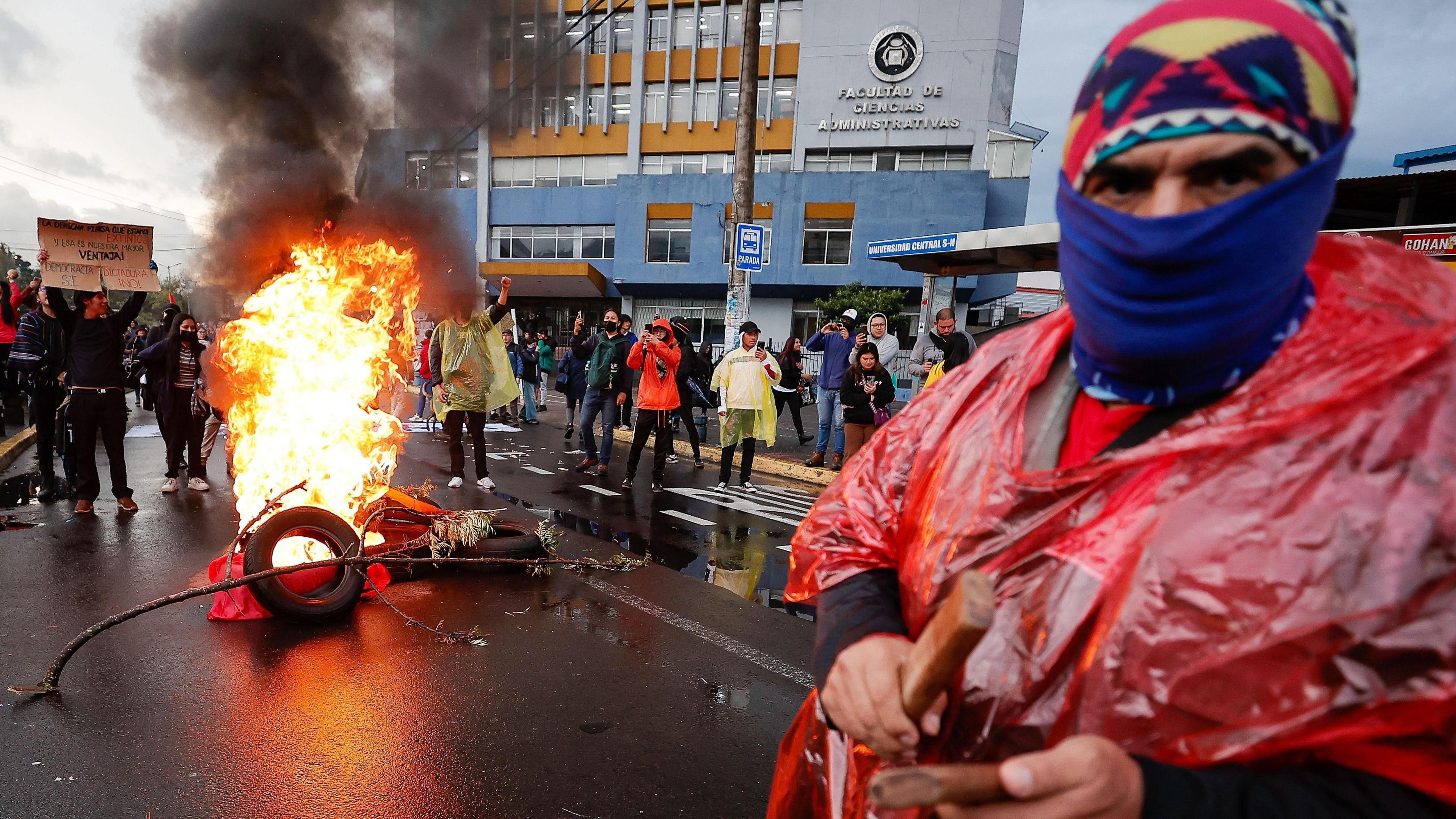 In the background a group gathers around a tire fire in the middle of a street, near a government building while in the foreground a man in a red poncho, blue balaklava and patterned red hat stares out at the camera