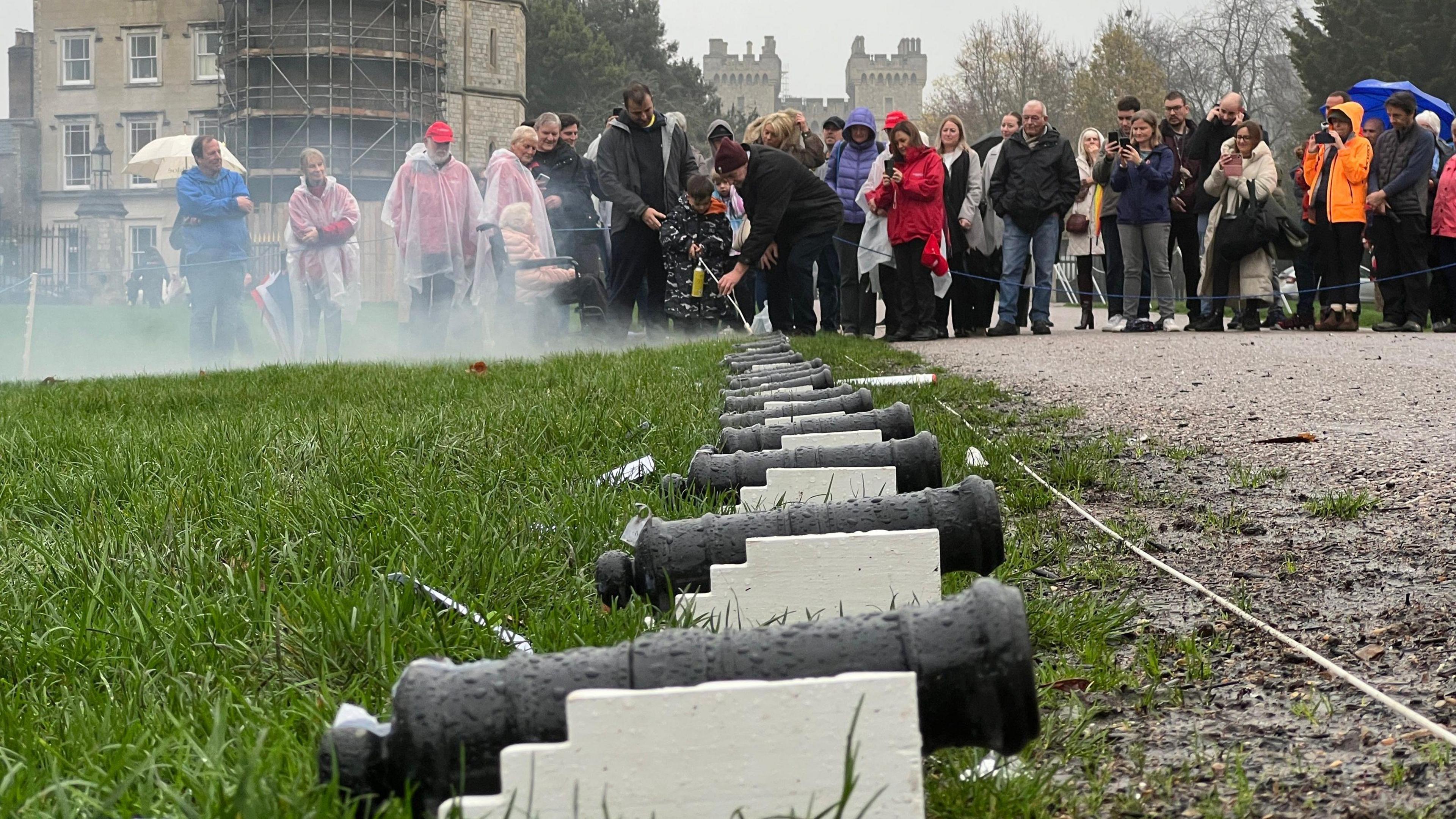 People stood in the rain with mini cannons laid out in front of them on a lawn.
