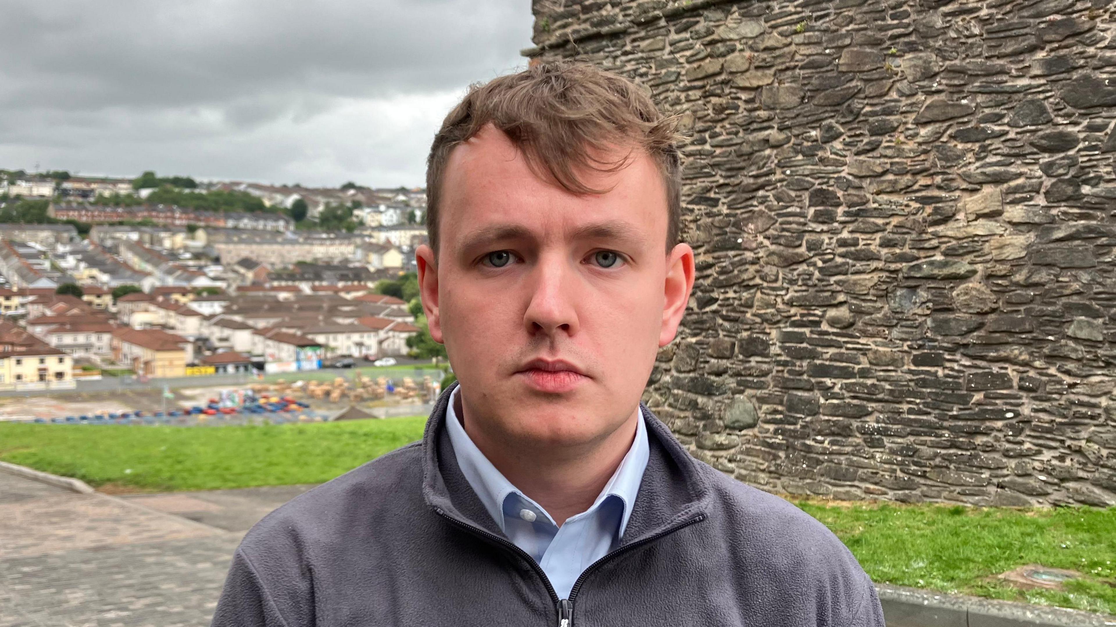 Pádraig Delargy has short brown hair. He is is wearing a grey fleece and a blue shirt. He is standing near to Derry's historic walls and a number of houses in the Bogside can be seen in the background.