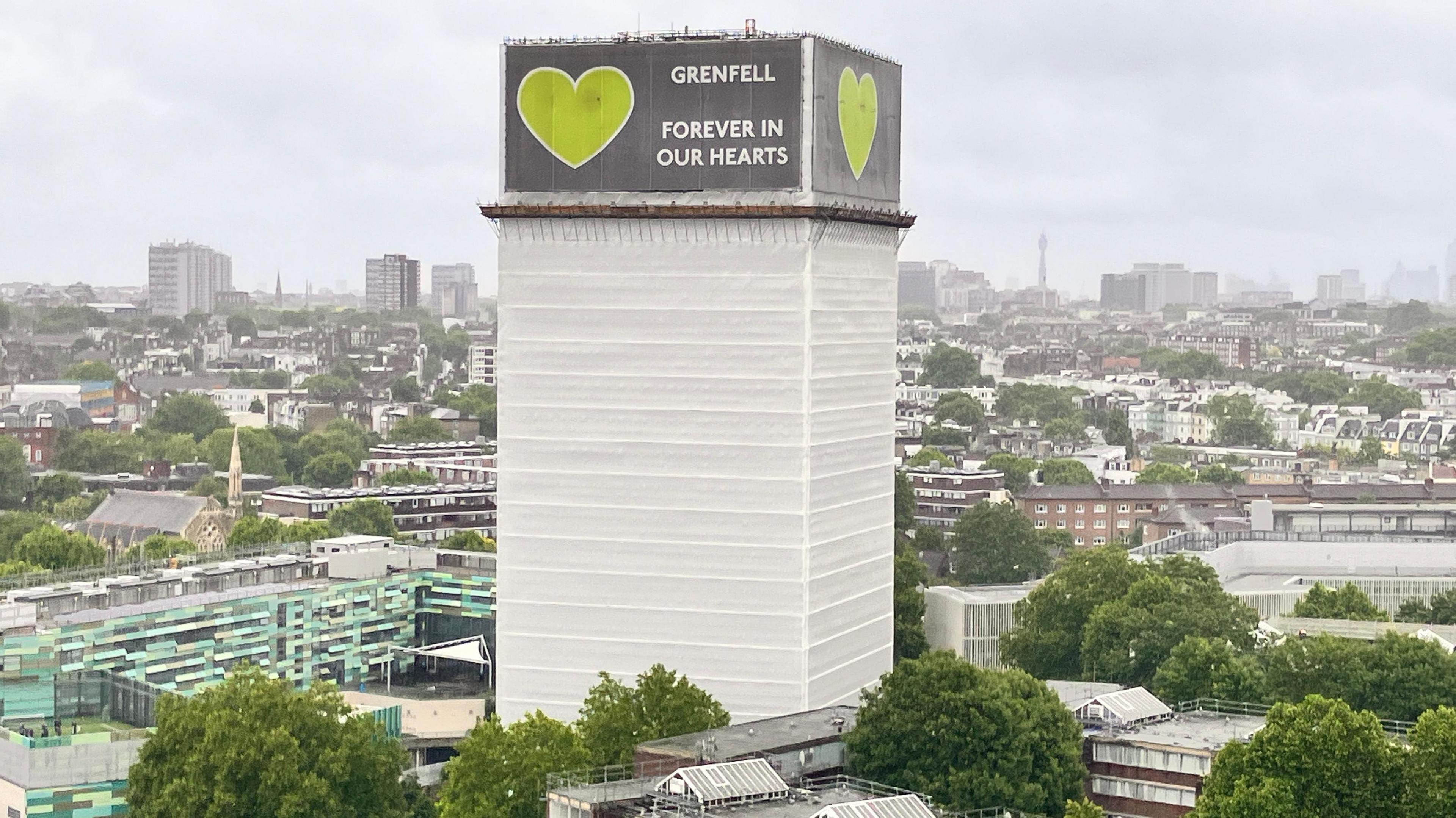 Grenfell tower which is covered in white hoardings and a green and grey sign around the top floors saying "GRENFELL FOREVER IN OUR HEARTS" and a green heart.