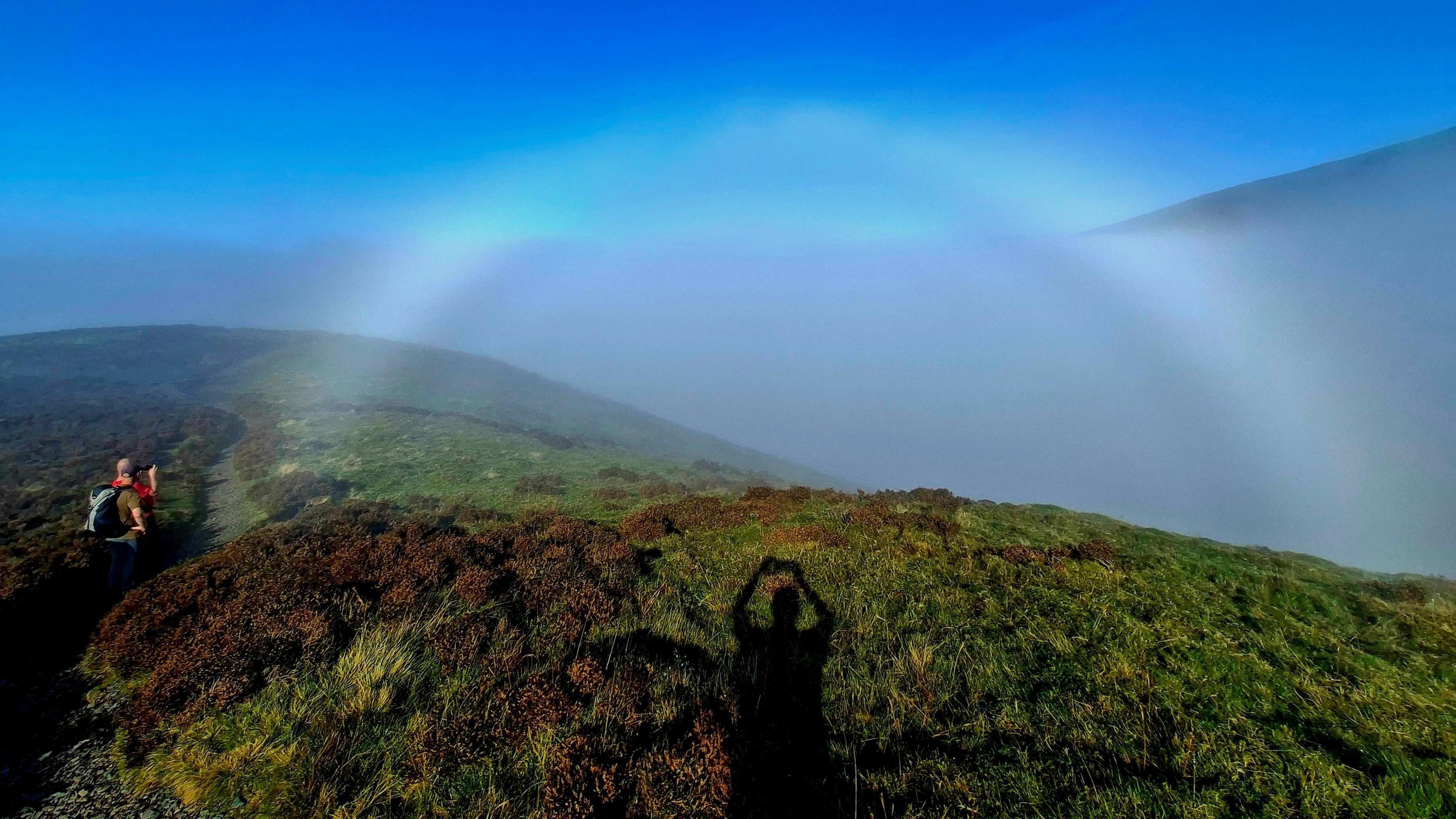 Sunlight and mist create the arch of a rainbow. There is shadow of a person holding a camera in the foreground, while two other people are off to the left, one of whom is also taking a photo. The landscape is rolling green hills with the outline of a larger mountain in the background, set against a vibrant blue sky.