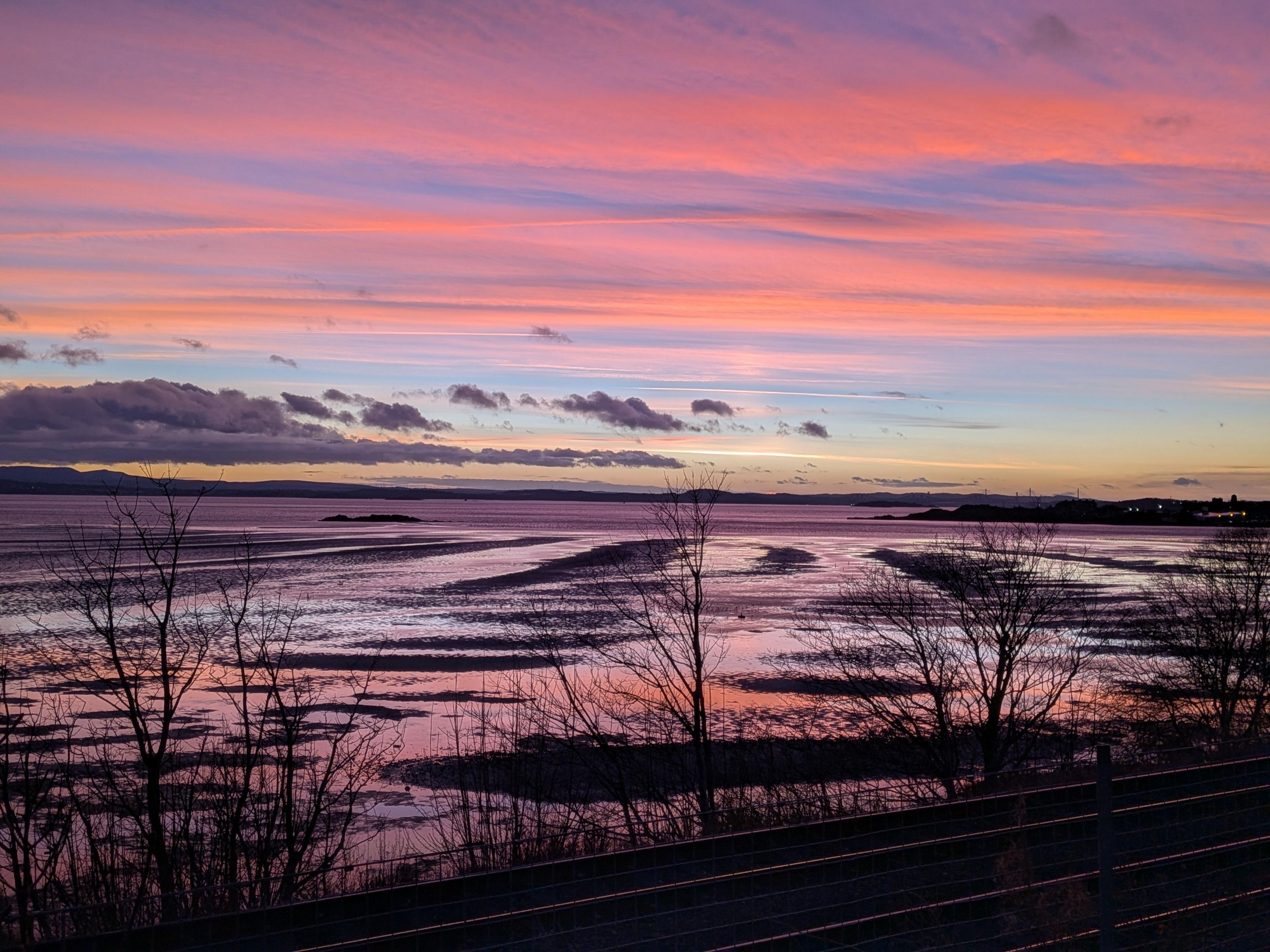 Sunset looking across a bay. The sky is different shades of pink and purple and is reflected on the water. There are also trees and clouds.