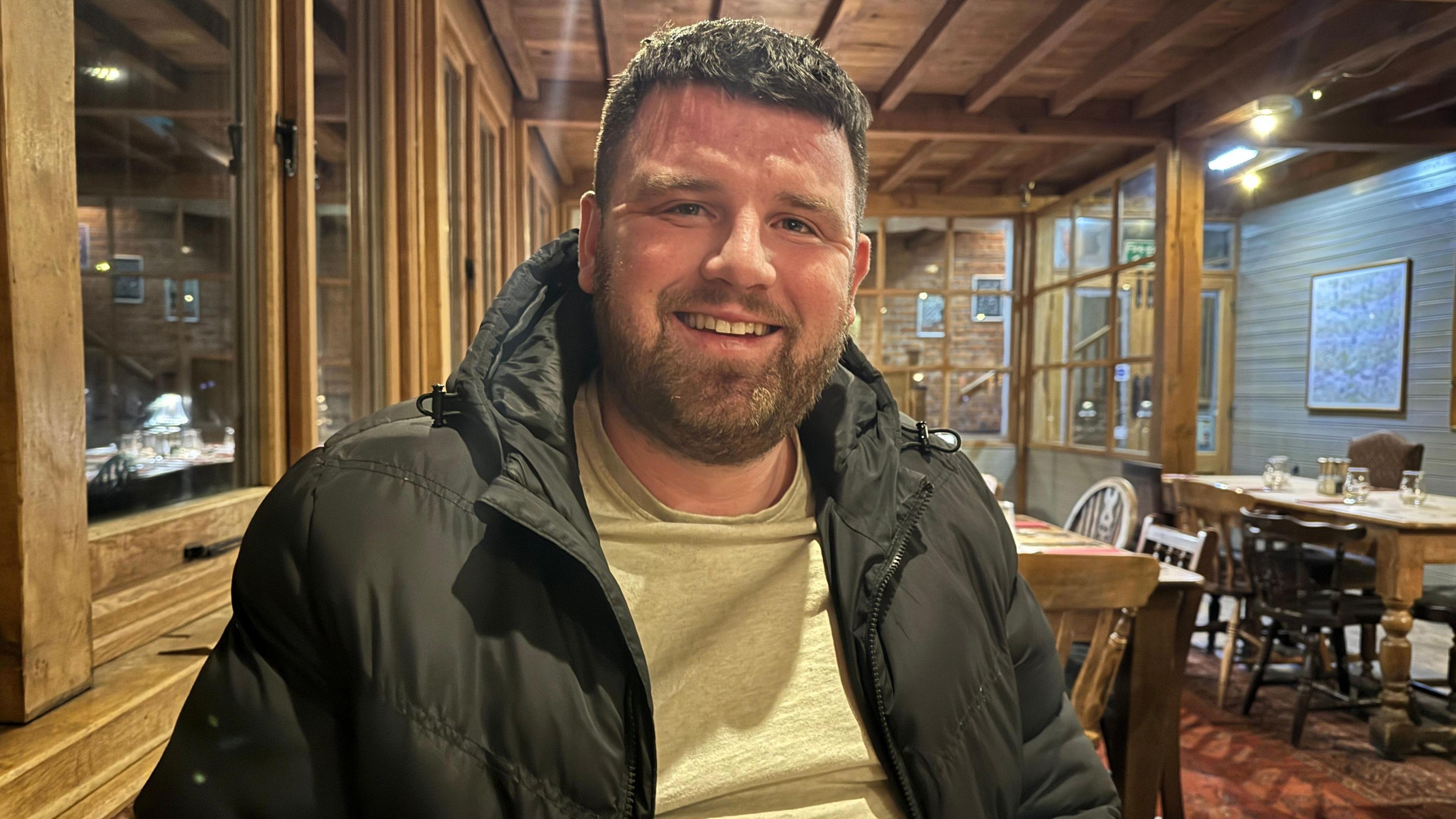 A man with brown hair and brown facial hair, sitting in a pub. He is wearing a grey t-shirt and black waterproof coat.