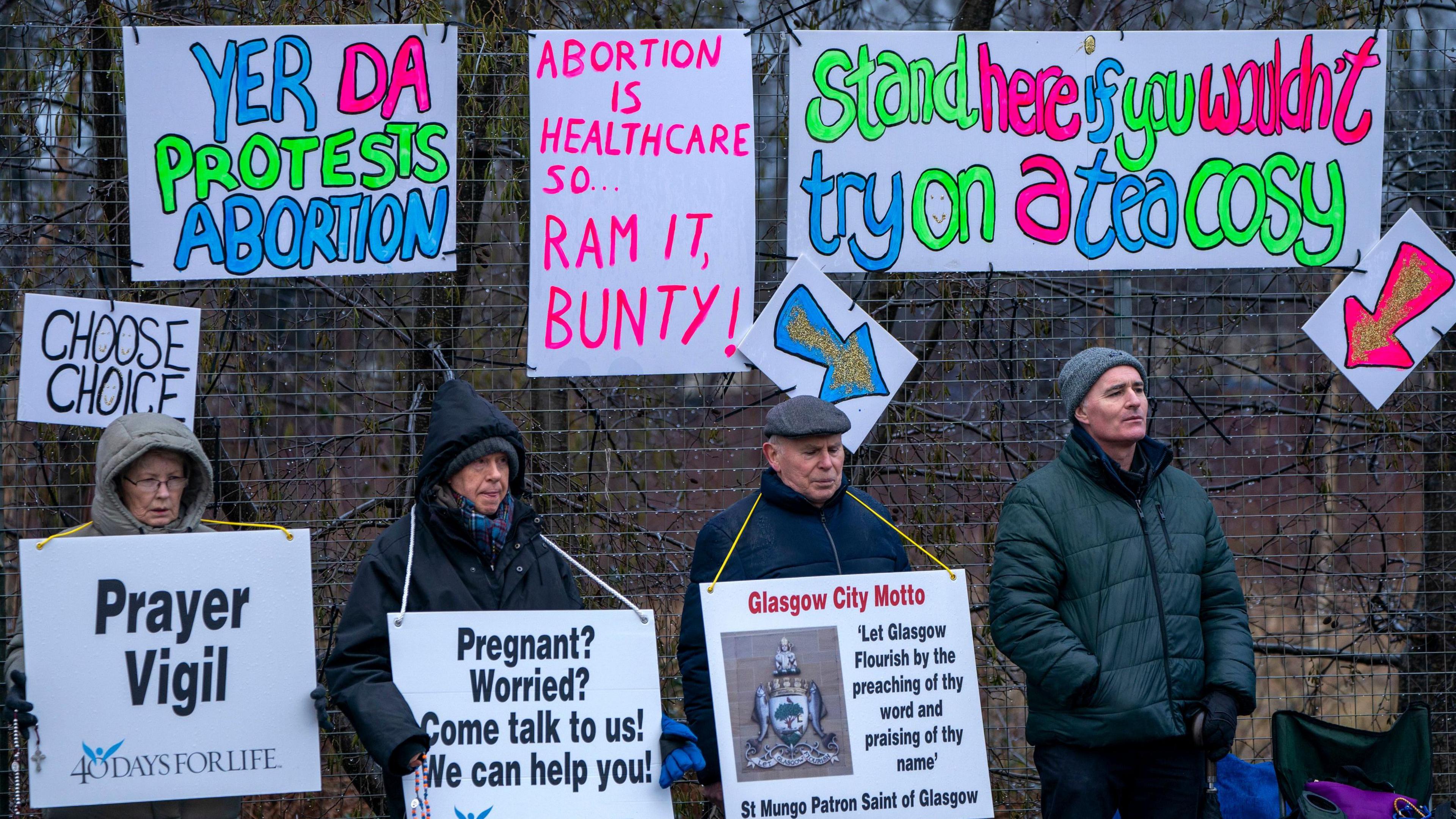 Pro-life protesters stand in zipped up coats and hats and hoods holding signs urging women to rethink abortion choices. Neon hand-made pro-choice signs in counter protest are hung on a fence behind them.