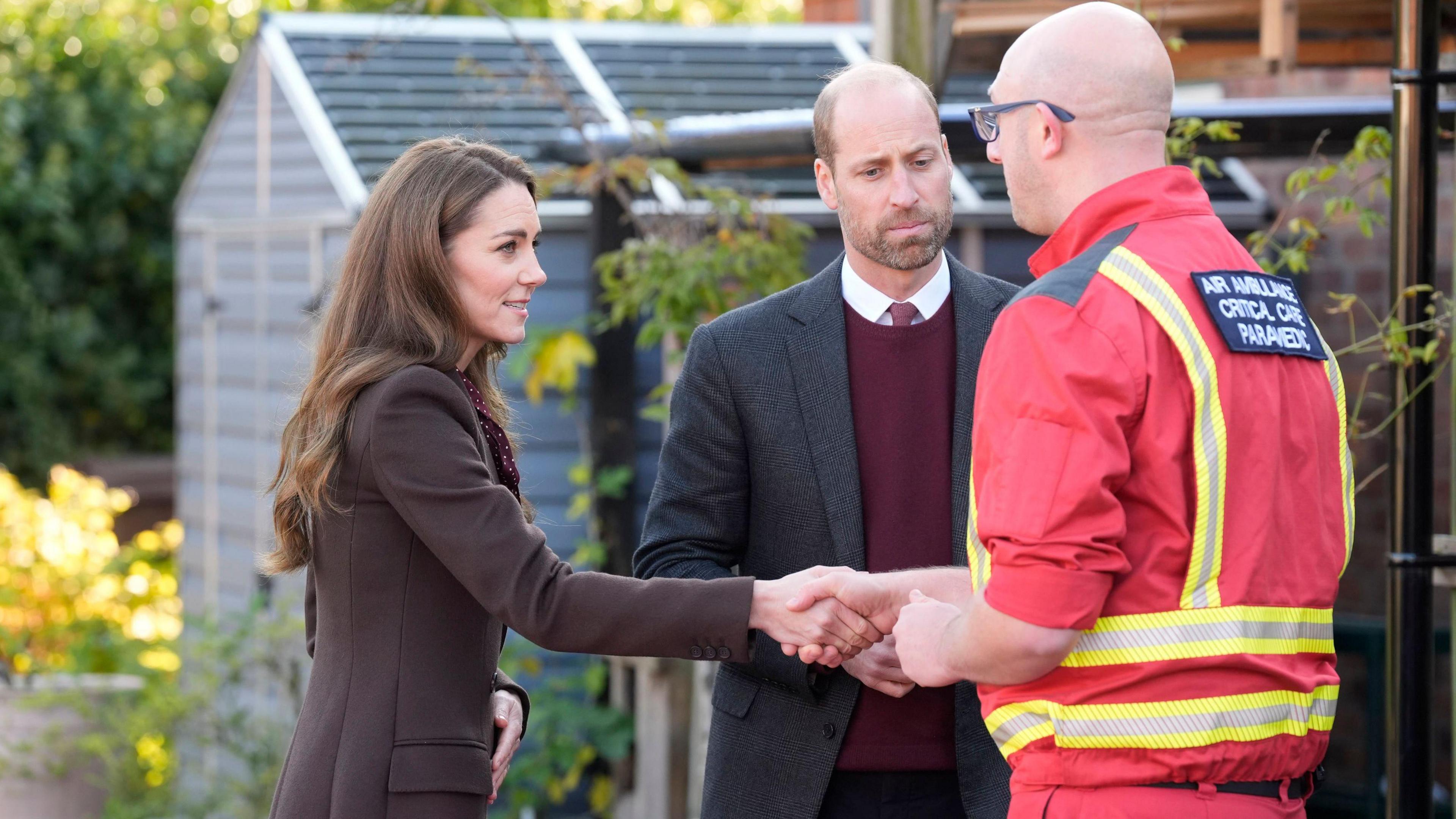 The Prince and Princess of Wales meet an air ambulance worker who responded to the Southport attacks. Princess Catherine is wearing a long brown coat and maroon blouse with polka dots. Prince William is wearing a maroon jumper and tie with a grey suit jacket, while the ambulance worker wears his red and hi vis uniform.